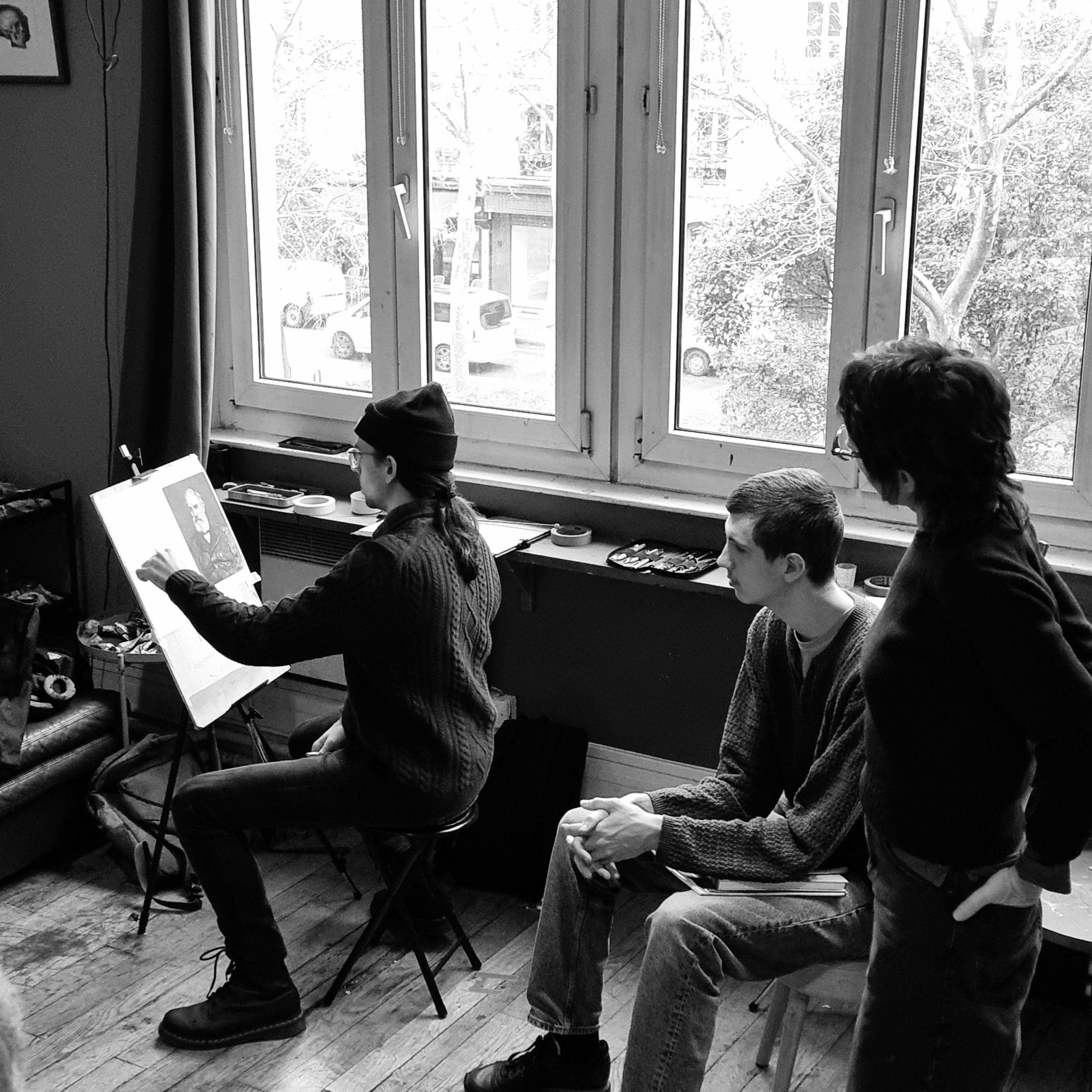 Three people are in an art studio. One person is sitting and drawing on an easel by a window, while two others look on. The room has wooden floors and art supplies on the windowsill.