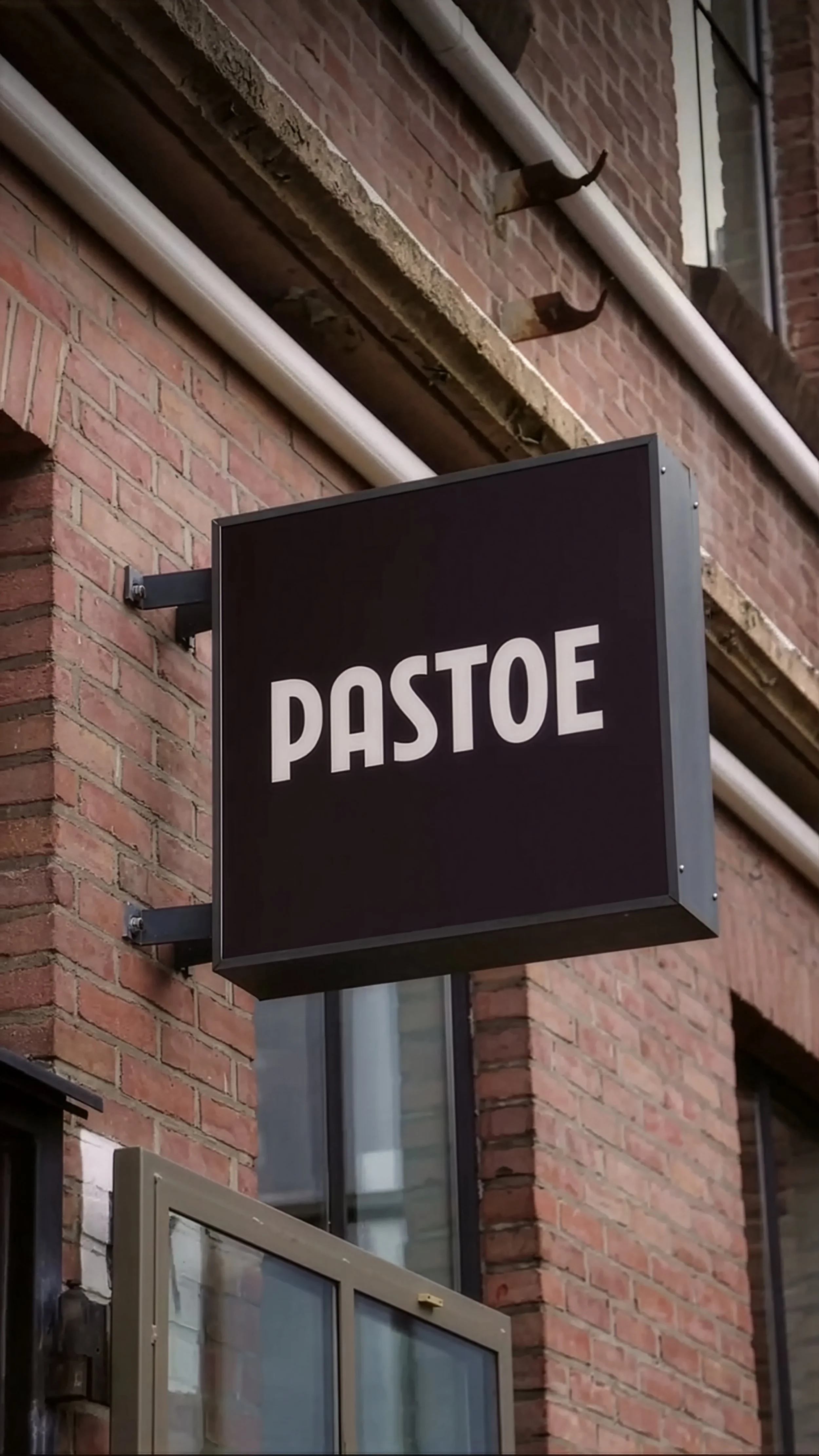 Close-up of a black "PASTOE" sign mounted on a red brick building, timeless dutch furniture design, branding and illustration project by Automne Studio.