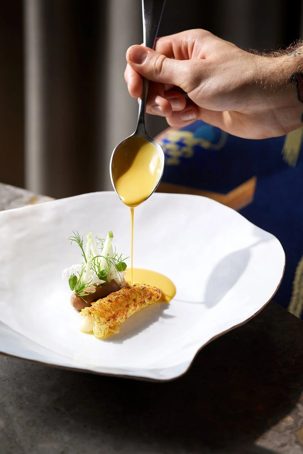 Chef drizzling yellow sauce from a spoon over a plated gourmet dish, Sarah-Linda, sensual tableware, branding, web design and editorial project by Automne Studio.