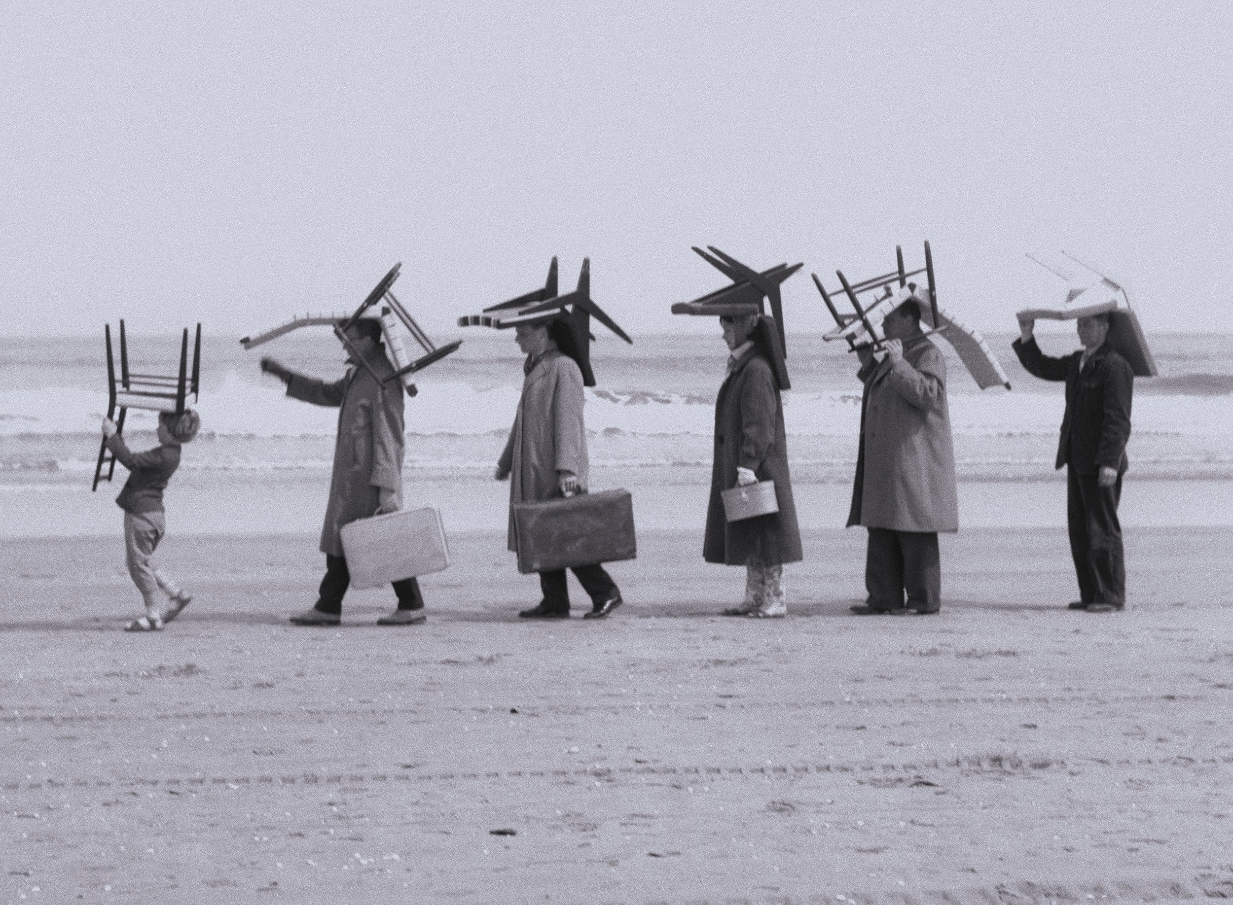 Black-and-white photo of six people walking along a beach carrying ladders, umbrellas, chairs, and briefcases, Pastoe, timeless dutch furniture design, branding project by Automne Studio.