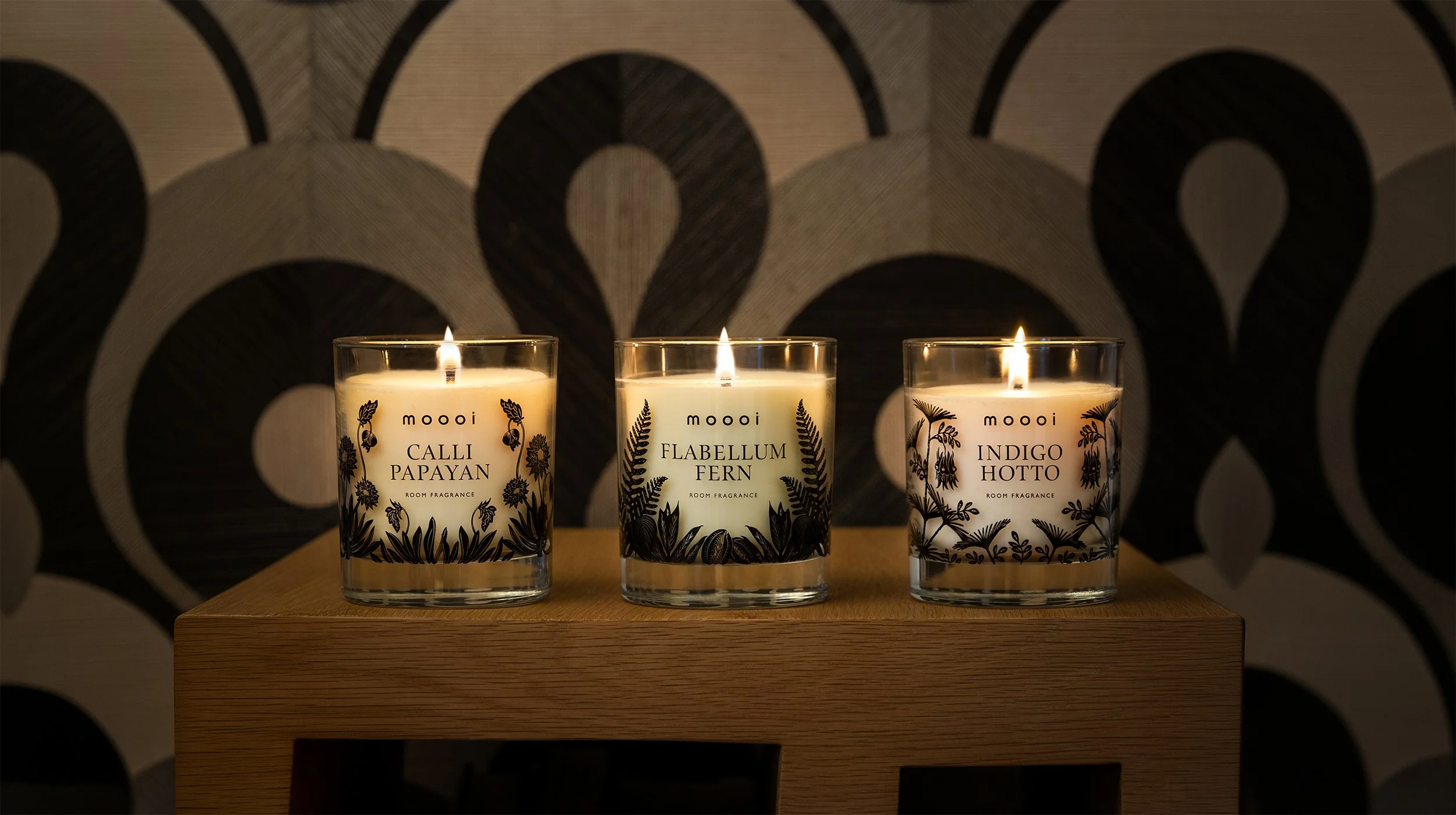 Three lit Moooi scented candles labeled Calli Papayan, Flabellum Fern, and Indigo Hotto, Extinct Plants Home Fragrance Collection by Moooi, illustration, packaging and visual identity by Automne Studio.