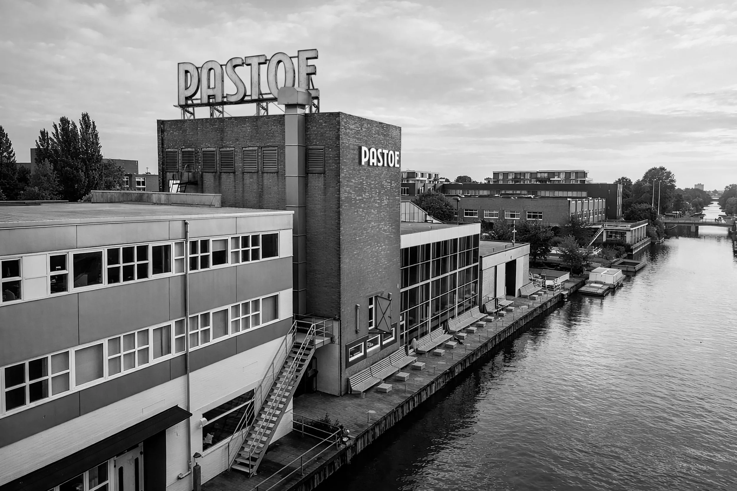 Black-and-white view of the waterfront industrial Pastoe building with large “PASTOE” rooftop signage, timeless dutch furniture design, branding and illustration project by Automne Studio.