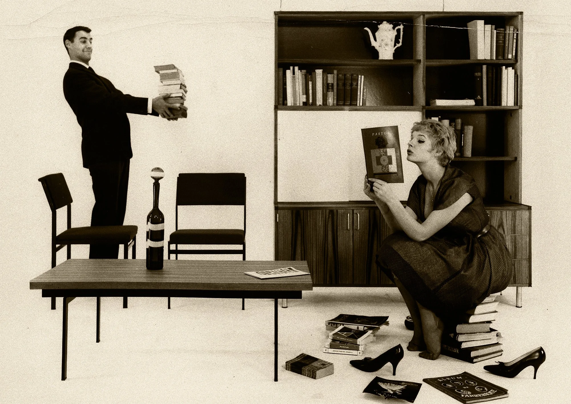 Vintage black-and-white photo of a man carrying books beside a table with wine, while a seated woman reads near a bookshelf, Pastoe, timeless dutch furniture design, branding and illustration project by Automne Studio.