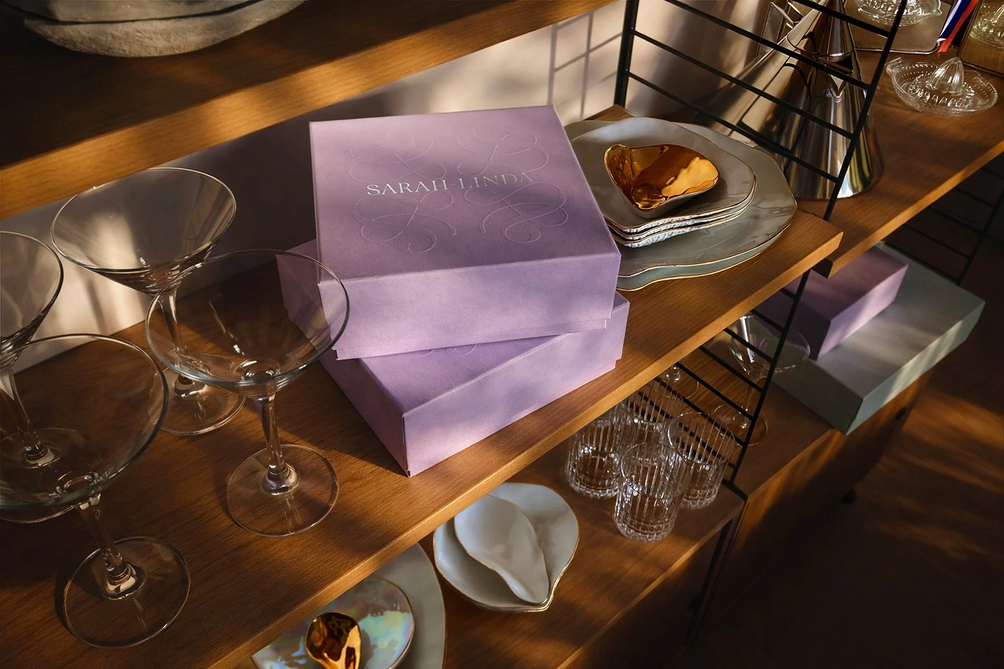 Purple Sarah Linda tableware boxes displayed on a wooden shelf with glassware, plates, and gold ceramic dishes, sensual tableware, branding, web design and editorial project by Automne Studio.