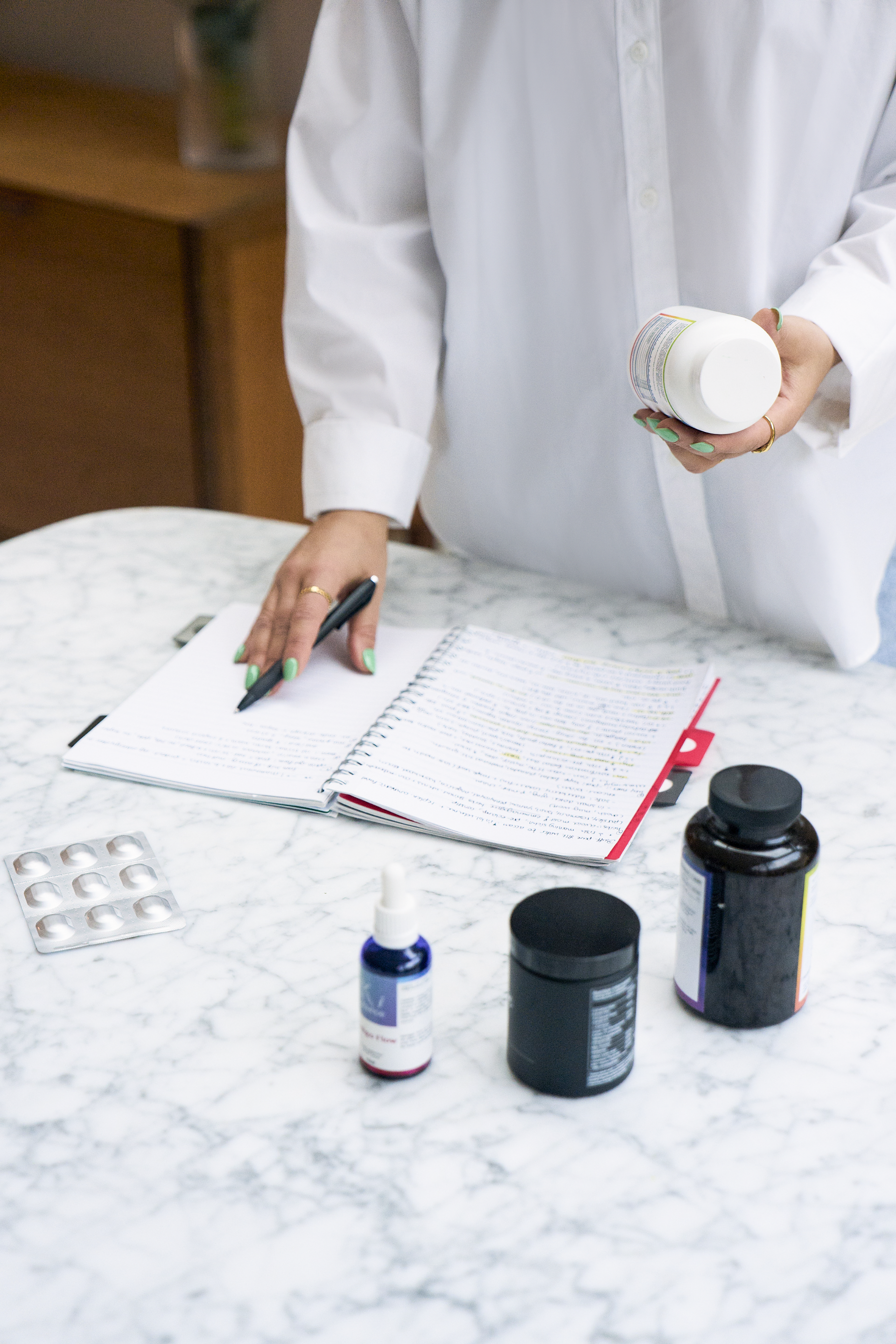 Person with green painted nails writing in a notebook on a marble table, surrounded by medication bottles and blister pack.