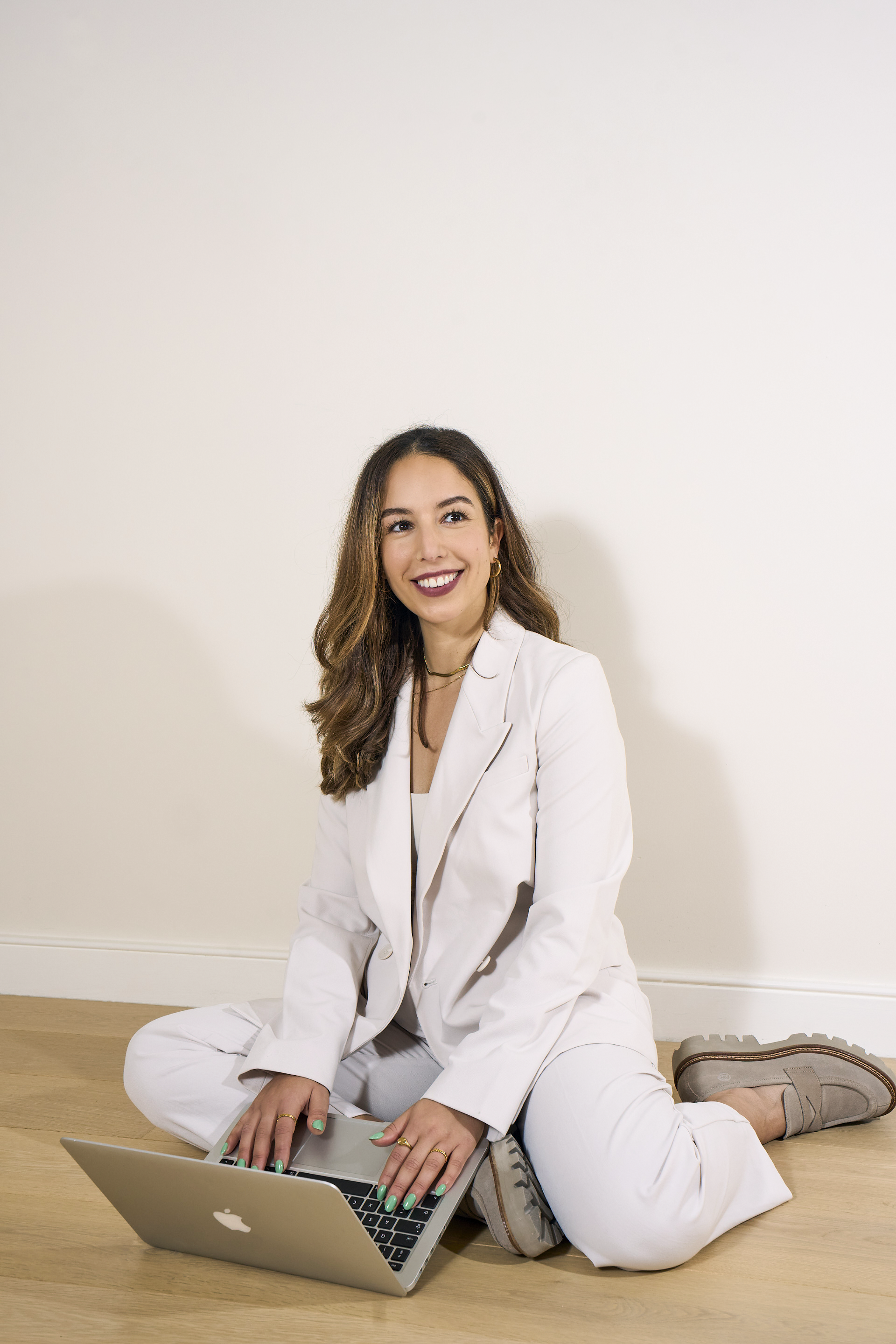 A woman sitting on a wooden floor with a laptop, wearing a white suit, smiling, and using the laptop.