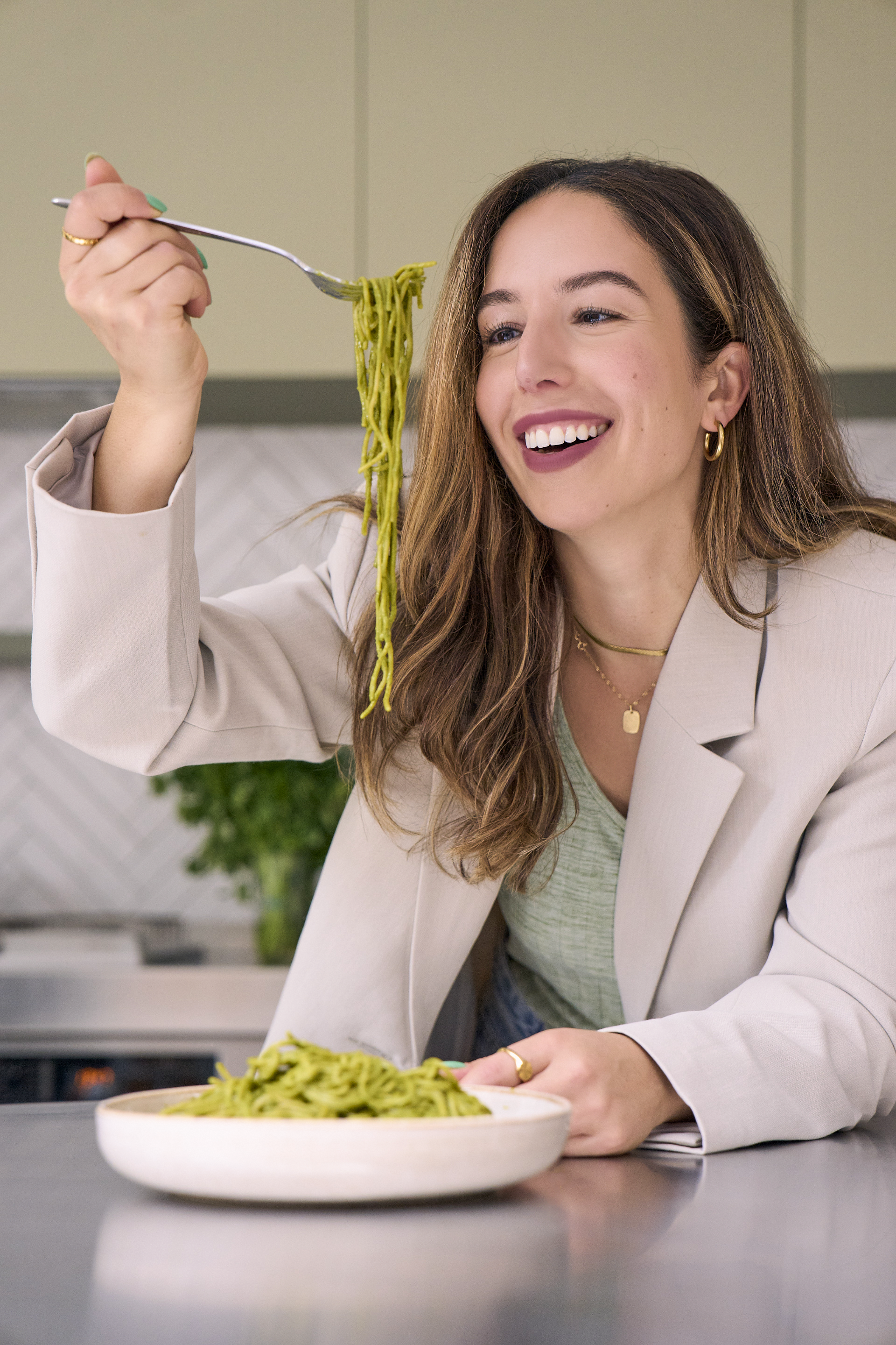 Woman in beige blazer holding fork with green pasta, smiling at plate of green pasta on countertop