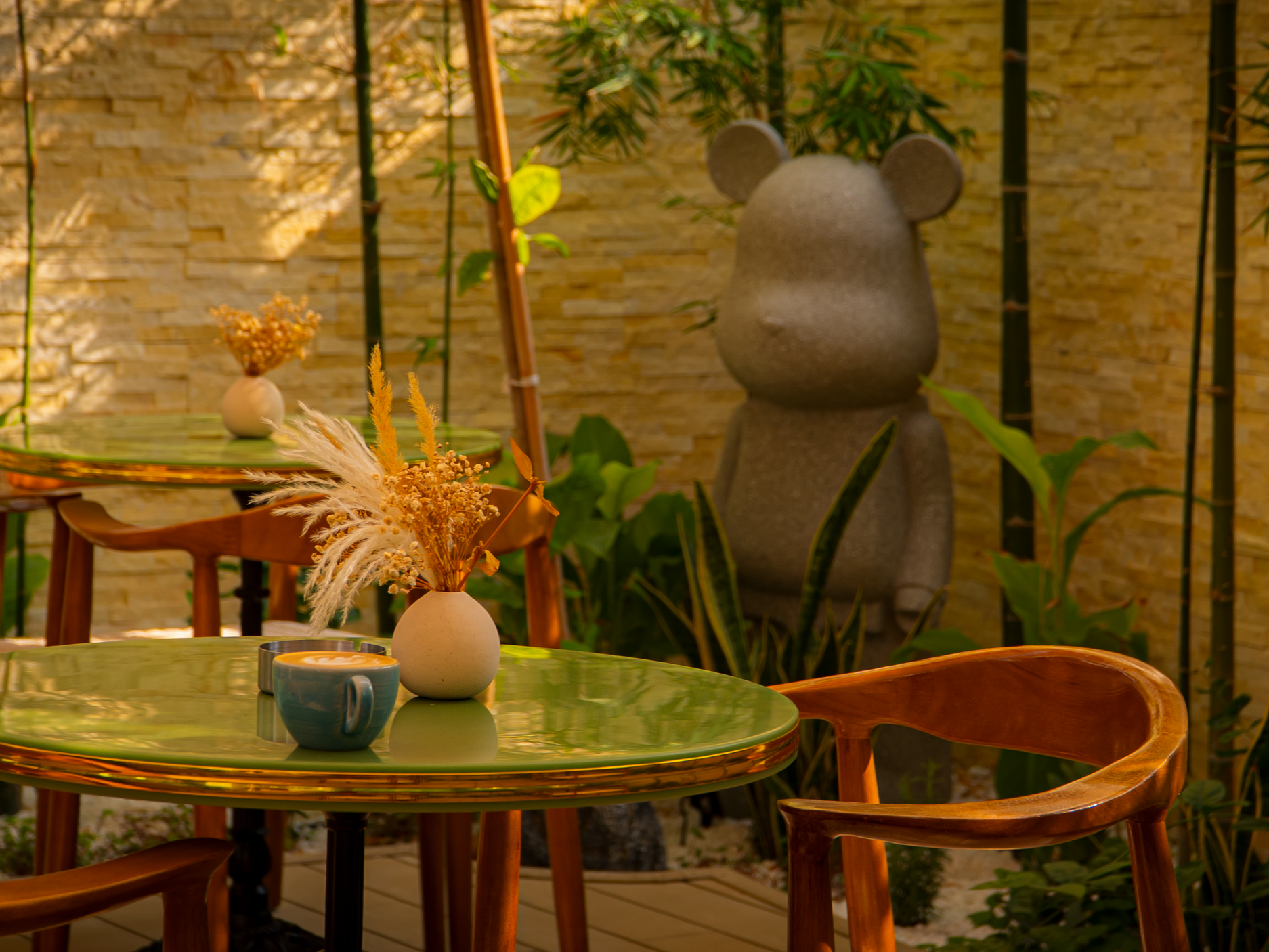 Indoor seating area with green glass table, wooden chairs, a vase with dried flowers, a coffee cup, and a stone sculpture of a bear with round ears surrounded by plants and a textured yellow wall.