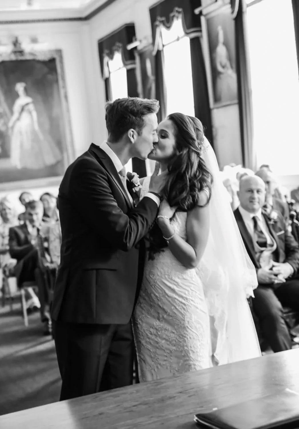 A black and white photo of a bride and groom kissing during their wedding ceremony in a decorated church, with seated guests watching.