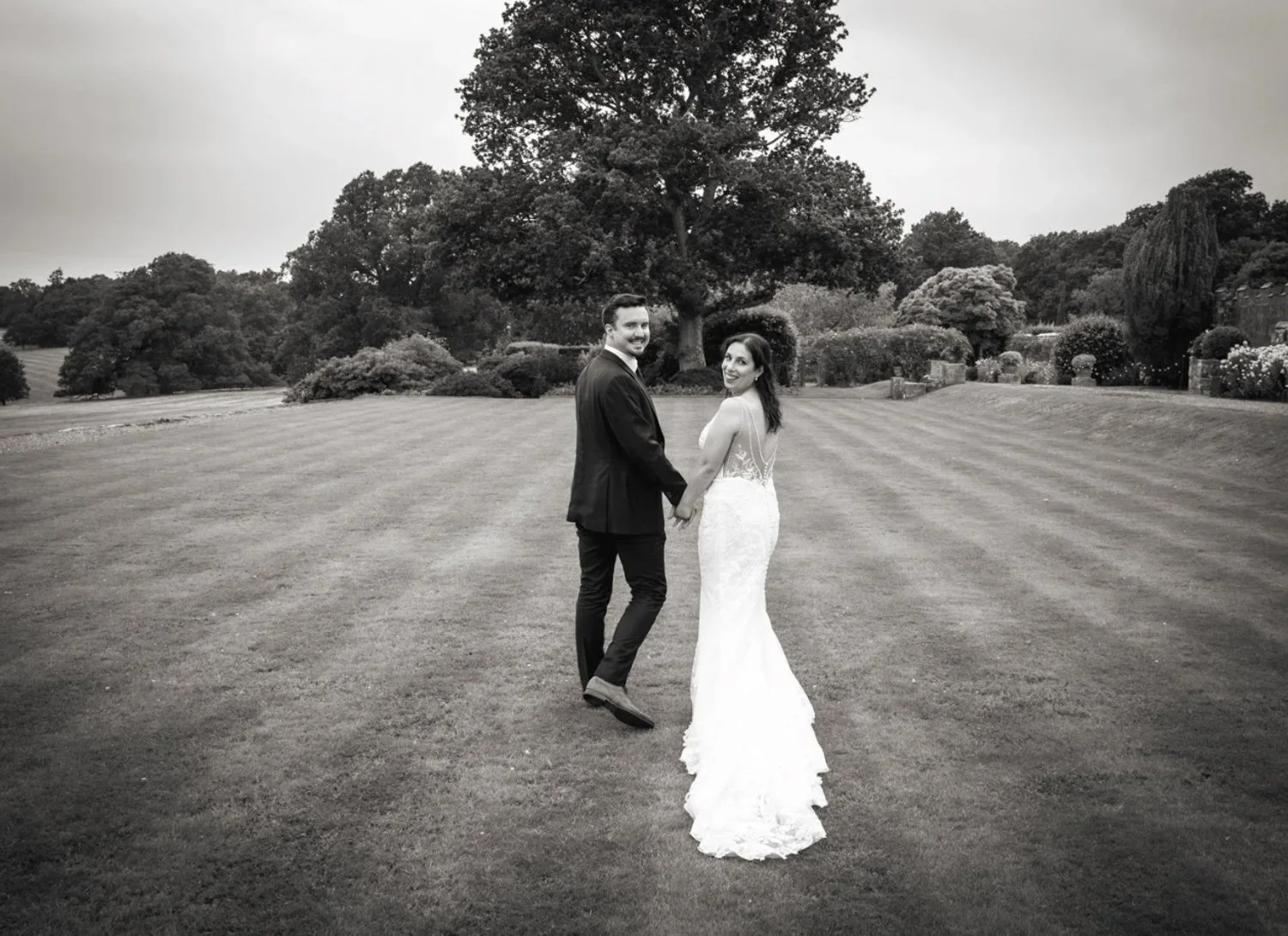 A bride and groom holding hands and facing the camera on a grassy field with trees and shrubs in the background.