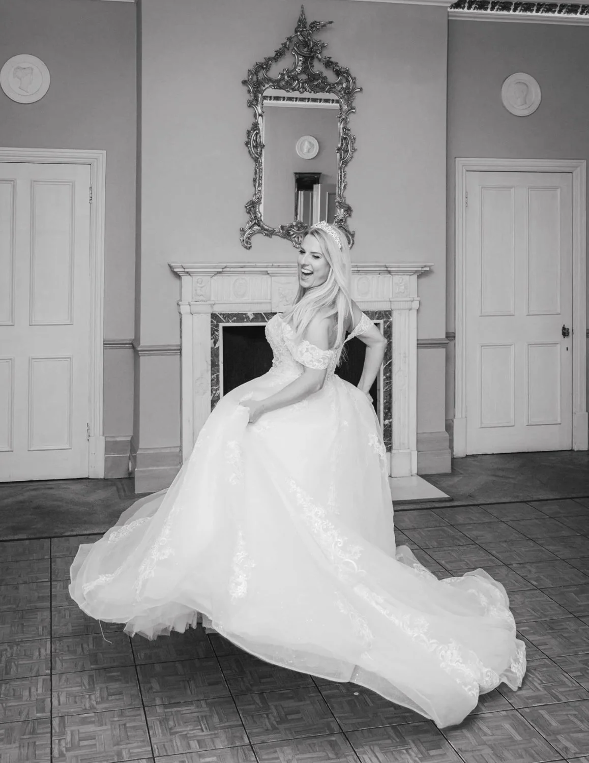 A smiling woman in a wedding dress sitting on her skirt in a room with a fireplace and ornate mirror.