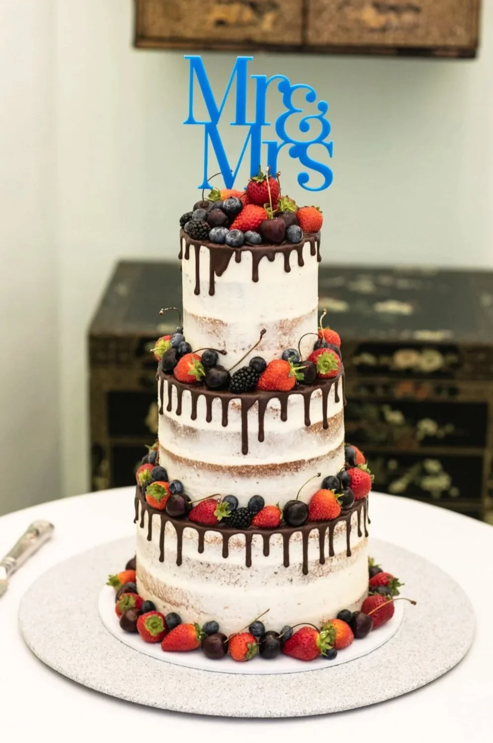 A three-tiered wedding cake decorated with fresh berries and a blue 'Mr. & Mrs.' topper.