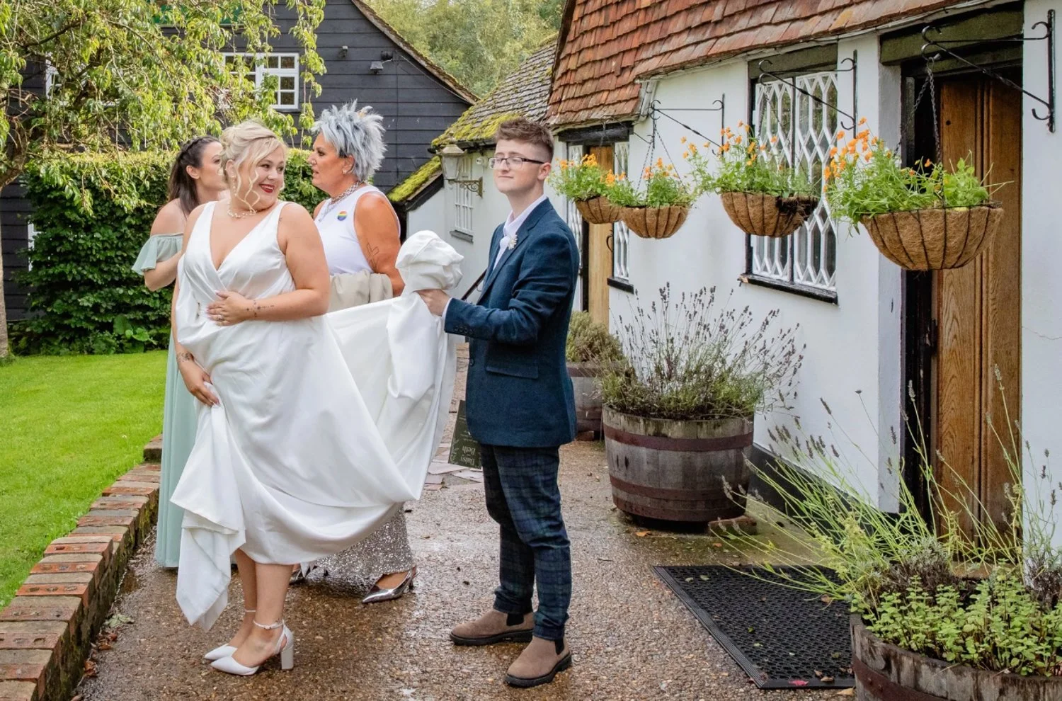 A bride in a white wedding dress is being carried by a woman in a pastel green dress, while a young man in a blazer and plaid pants holds her in a garden outside a cottage with hanging flower baskets and outdoor plants.