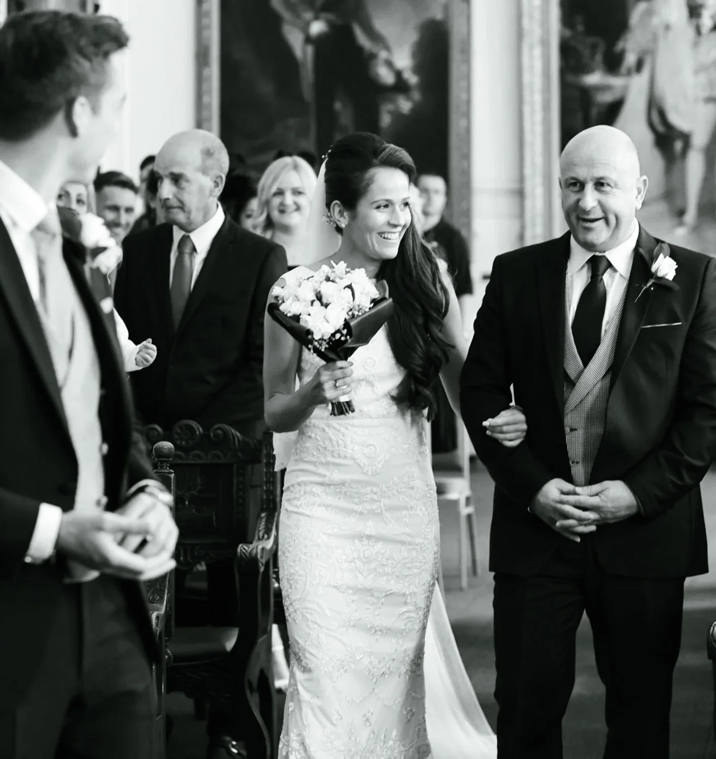 A bride smiling with a bouquet of flowers walking down the aisle with her father at her wedding ceremony, surrounded by celebrating guests.