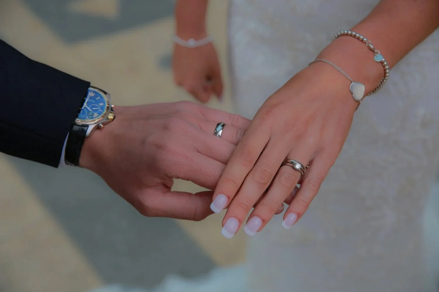 The bride and groom are holding hands, showing rings and bracelets. The groom wears a black suit with a blue watch, and the bride has manicured nails, a ring, and silver jewelry.