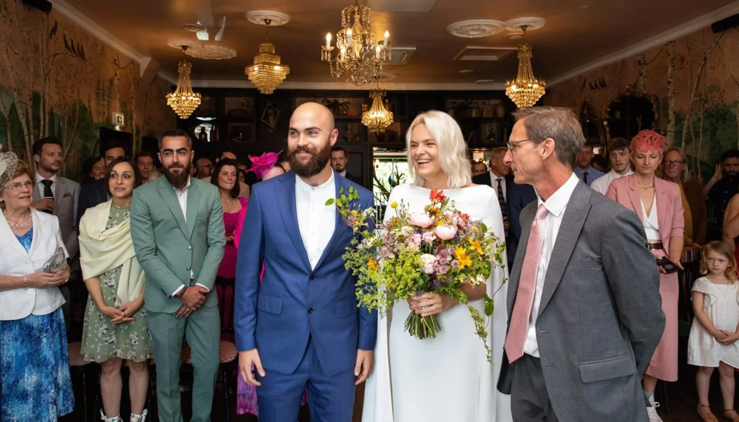 A wedding ceremony with the bride and groom standing with two older individuals, possibly parents, in a decorated indoor venue. The bride is holding a large bouquet of flowers and has blonde hair. The groom has a beard and is wearing a blue suit. The