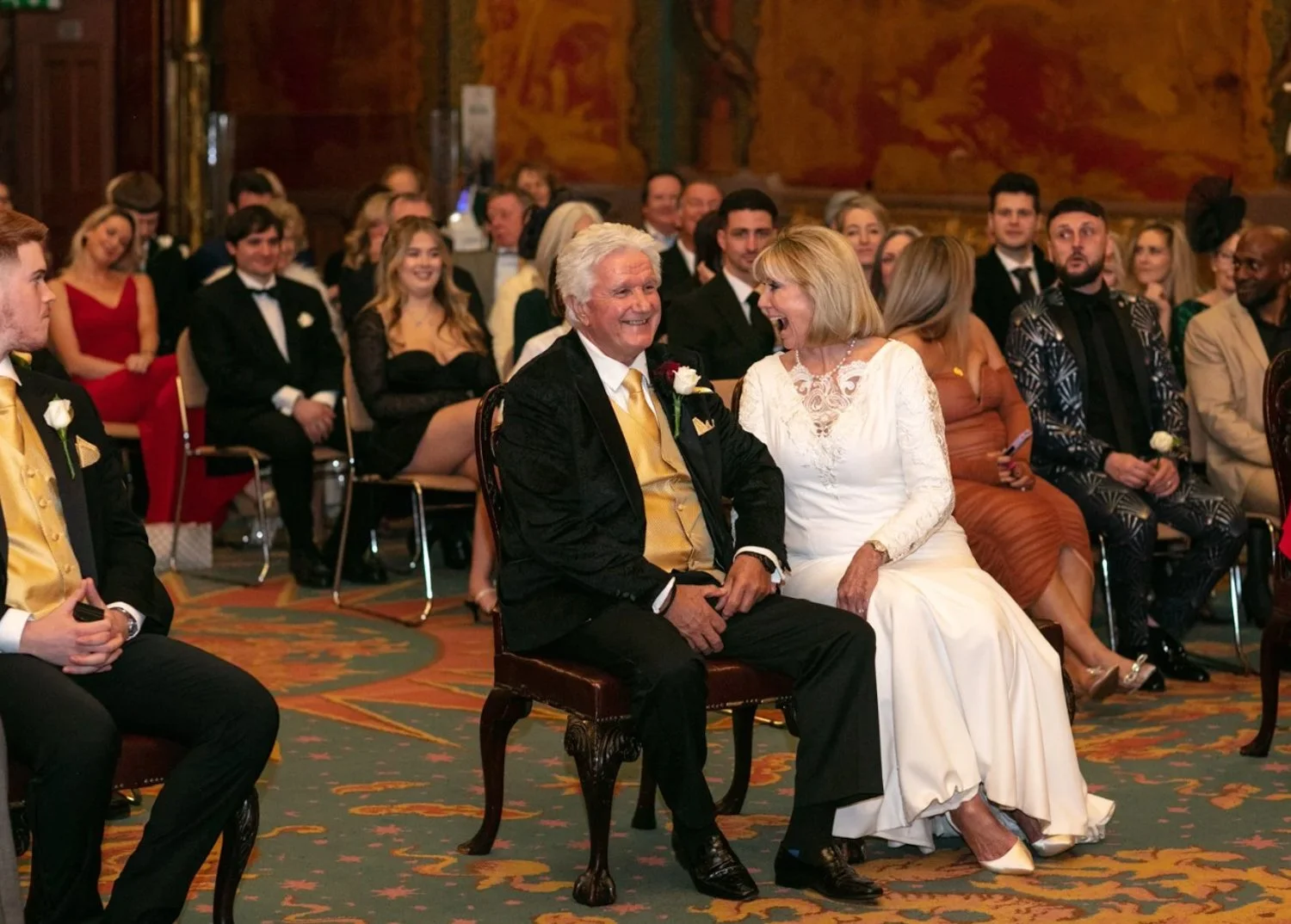 A group of people attending a formal event or wedding, with two people in the foreground sitting on an ornate chair, smiling and engaging in conversation. The woman is wearing a white gown, and the man is dressed in a black tuxedo with a gold vest an