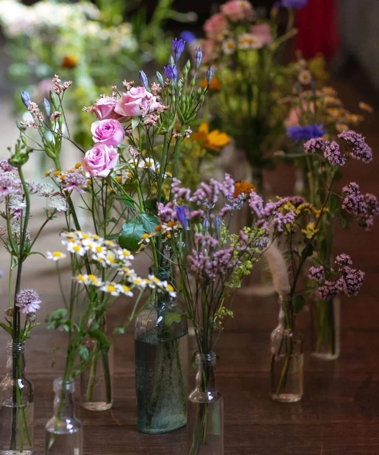 Multiple glass bottles of various sizes and colors filled with assorted colorful wildflowers on a wooden surface.