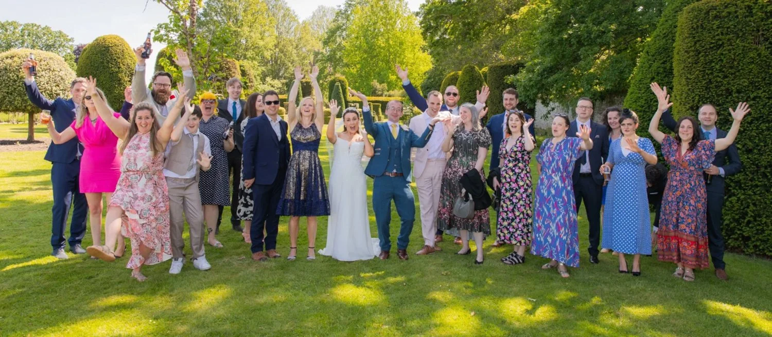 Group of people celebrating outdoors on a sunny day, dressed in formal and casual attire, with some raising their hands and holding drinks, in a lush garden setting.