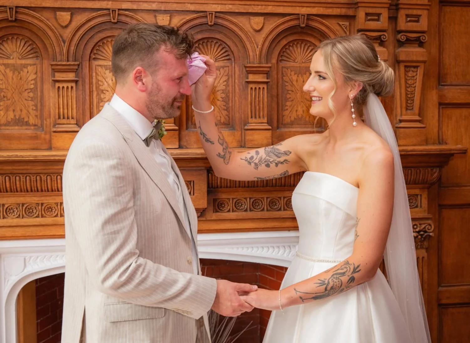 A bride and groom holding hands during their wedding ceremony, with the bride wiping away a tear from the groom's face in a wood-paneled room.