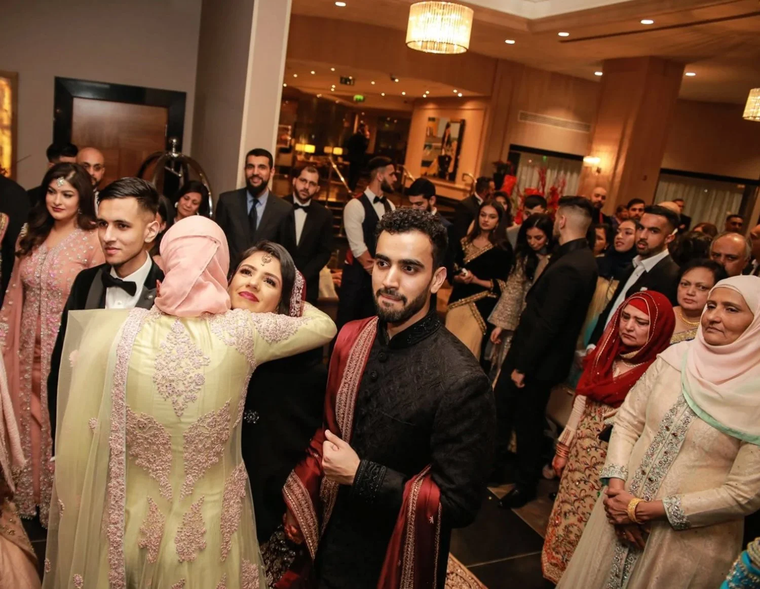 A group of people at a celebration or wedding, with a woman hugging a man in traditional South Asian attire and others dressed in formal wear, standing indoors with warm lighting.
