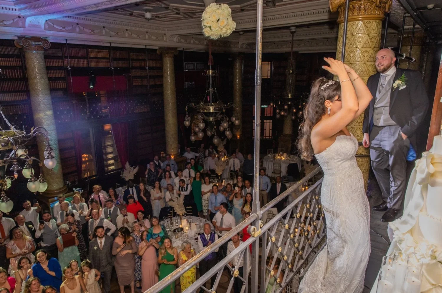 Bride and groom on a balcony during their wedding reception, with guests sitting at tables below in an elegant, decorated hall.
