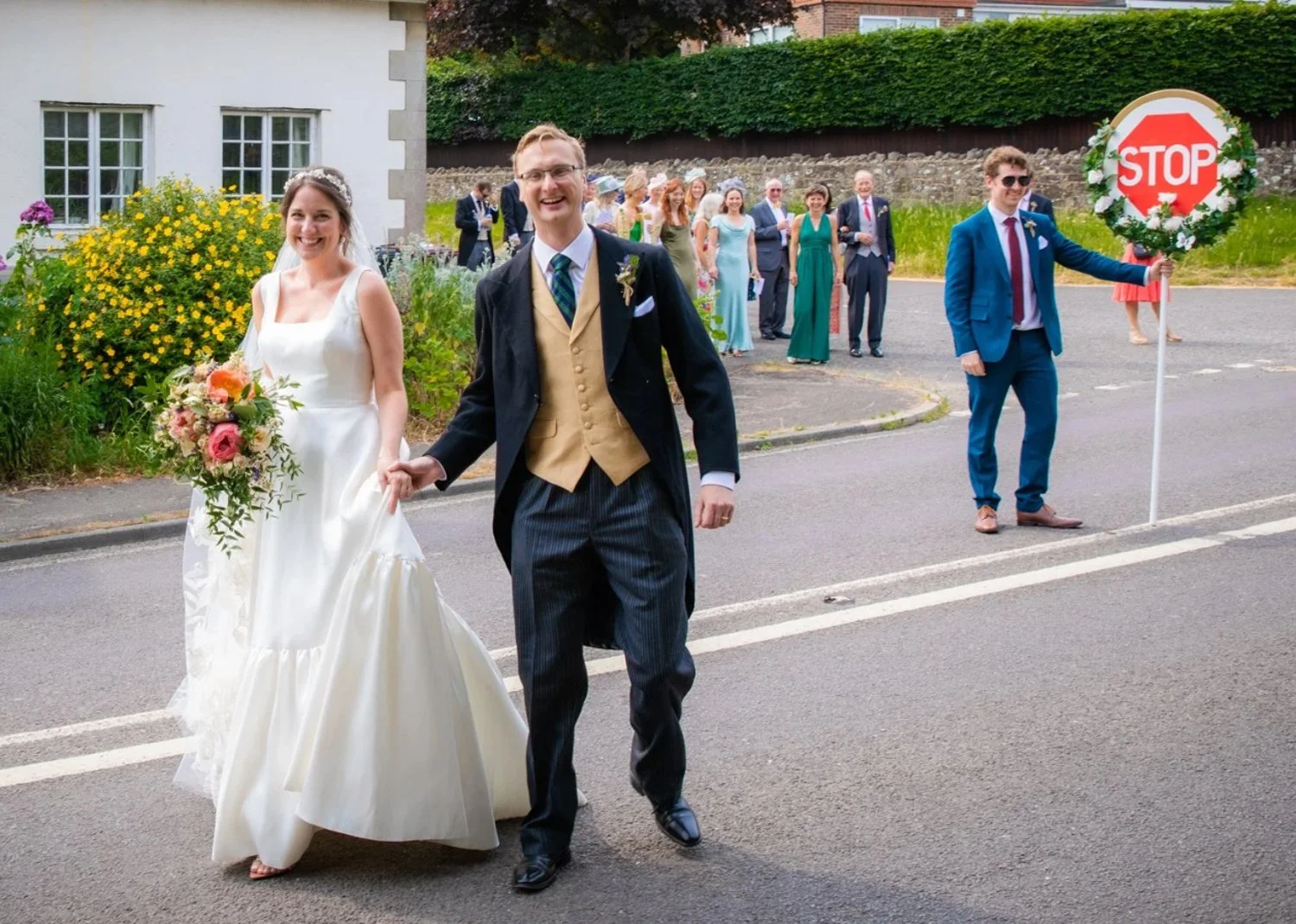 A wedding procession with a bride and groom walking hand in hand in the street, surrounded by friends and family. The bride is wearing a white wedding dress and holding a bouquet, while the groom is dressed in a formal suit with a vest and tie. Some