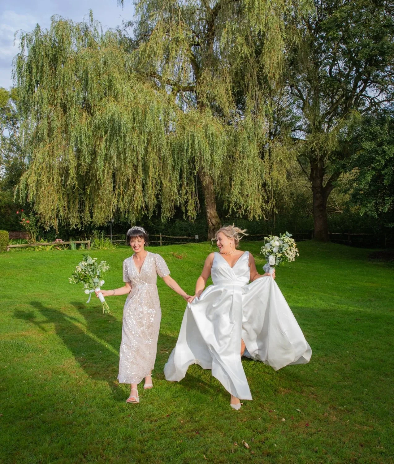 Two women in wedding dresses walking on green grass in a park, holding bouquets, with large trees in the background.