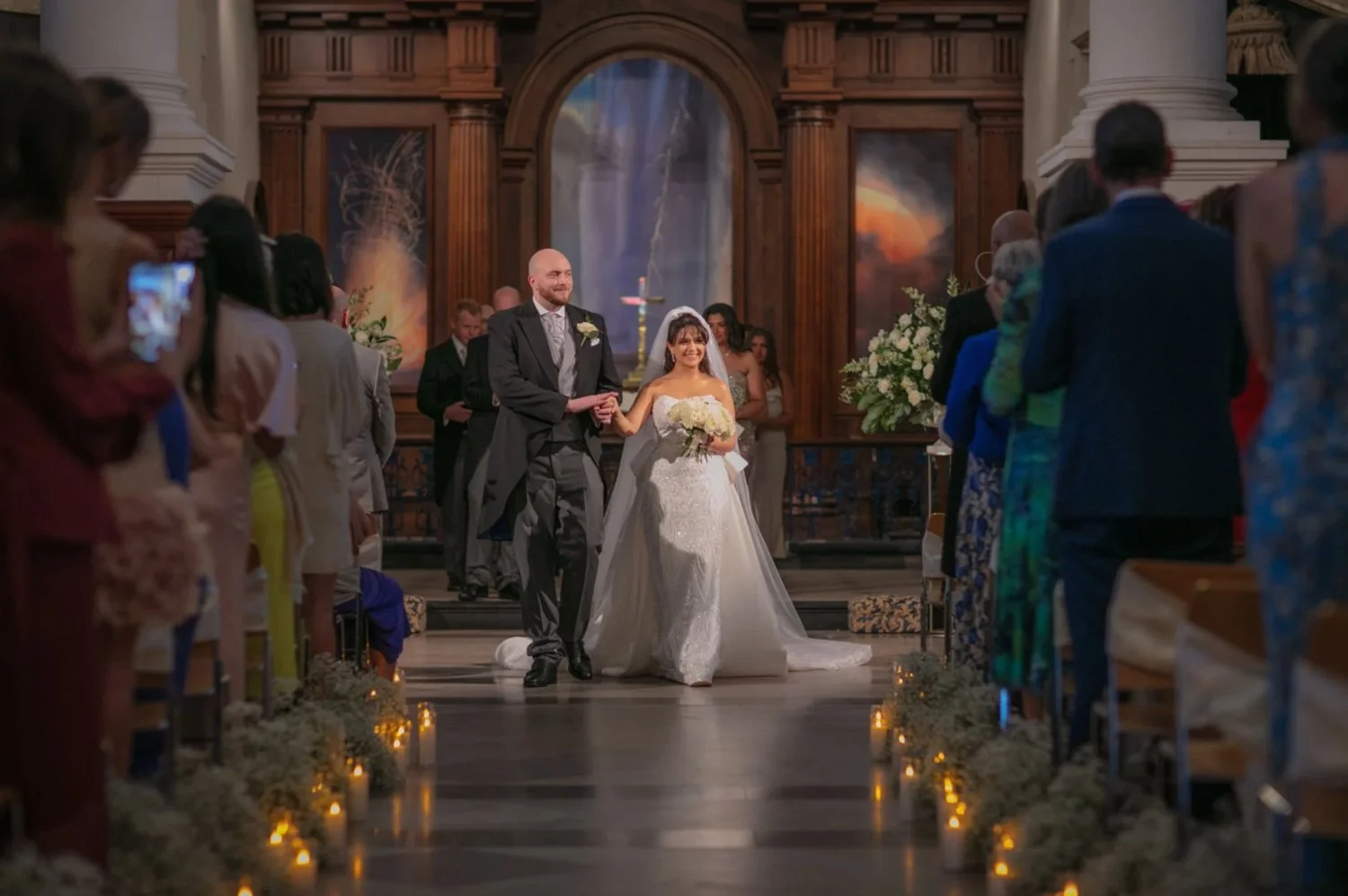 A couple of bride and groom walking down the aisle after their wedding ceremony inside a church, surrounded by seated wedding guests, with candles and floral arrangements along the aisle.