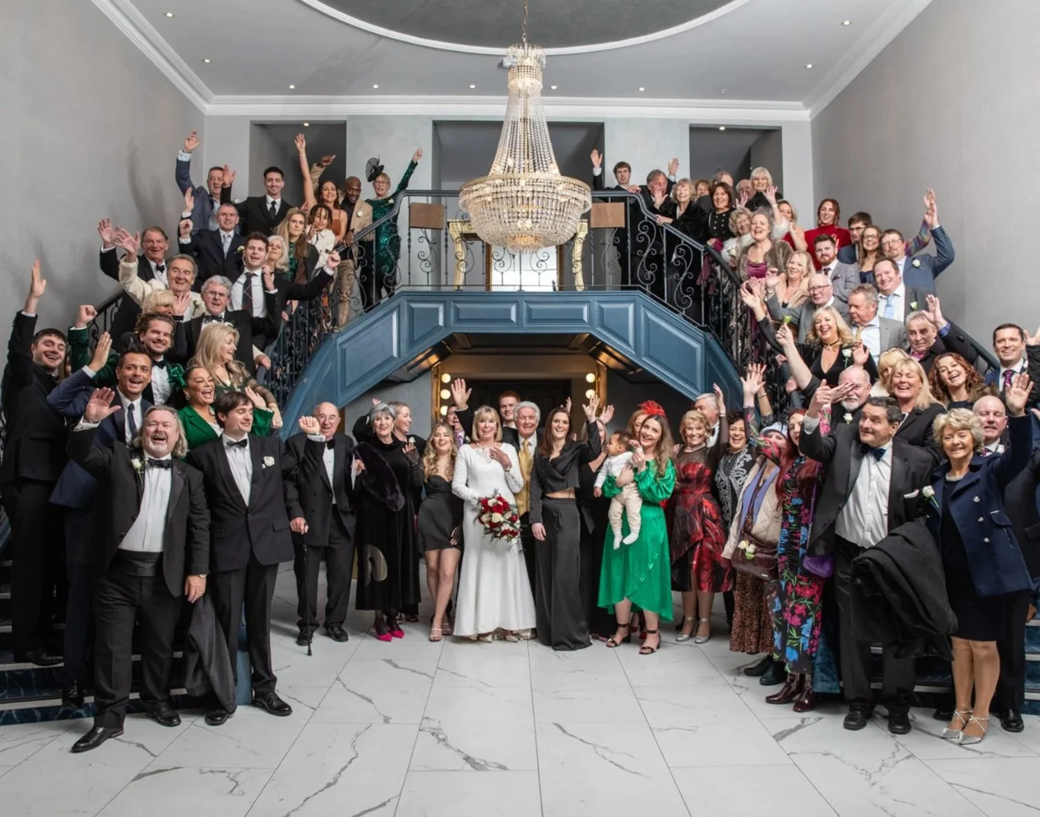 A large group of people dressed in formal attire, gathered inside a grand hall with a chandelier and a staircase, celebrating a wedding.
