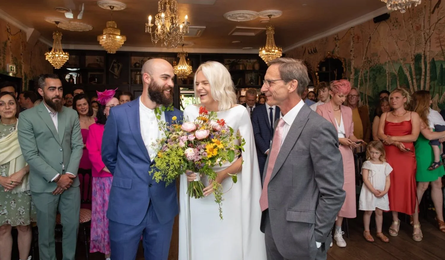 A wedding ceremony with a bride and groom smiling and holding a bouquet of flowers, surrounded by guests in formal attire in an ornately decorated room.