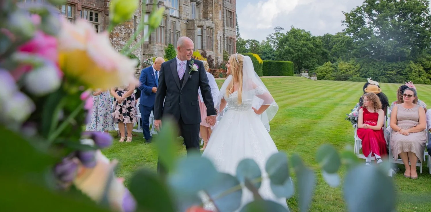 A bride and groom holding hands and smiling during their wedding ceremony outdoors, with guests seated on chairs around them, a historic building in the background, and floral decorations in the foreground.