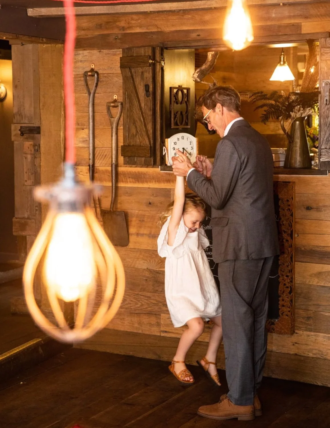A man and a young girl dance together in a cozy, rustic room with wooden walls. The man is wearing a gray suit and glasses, holding a pocket watch, while the girl, dressed in a white dress and tan sandals, is smiling and holding the man's hand above