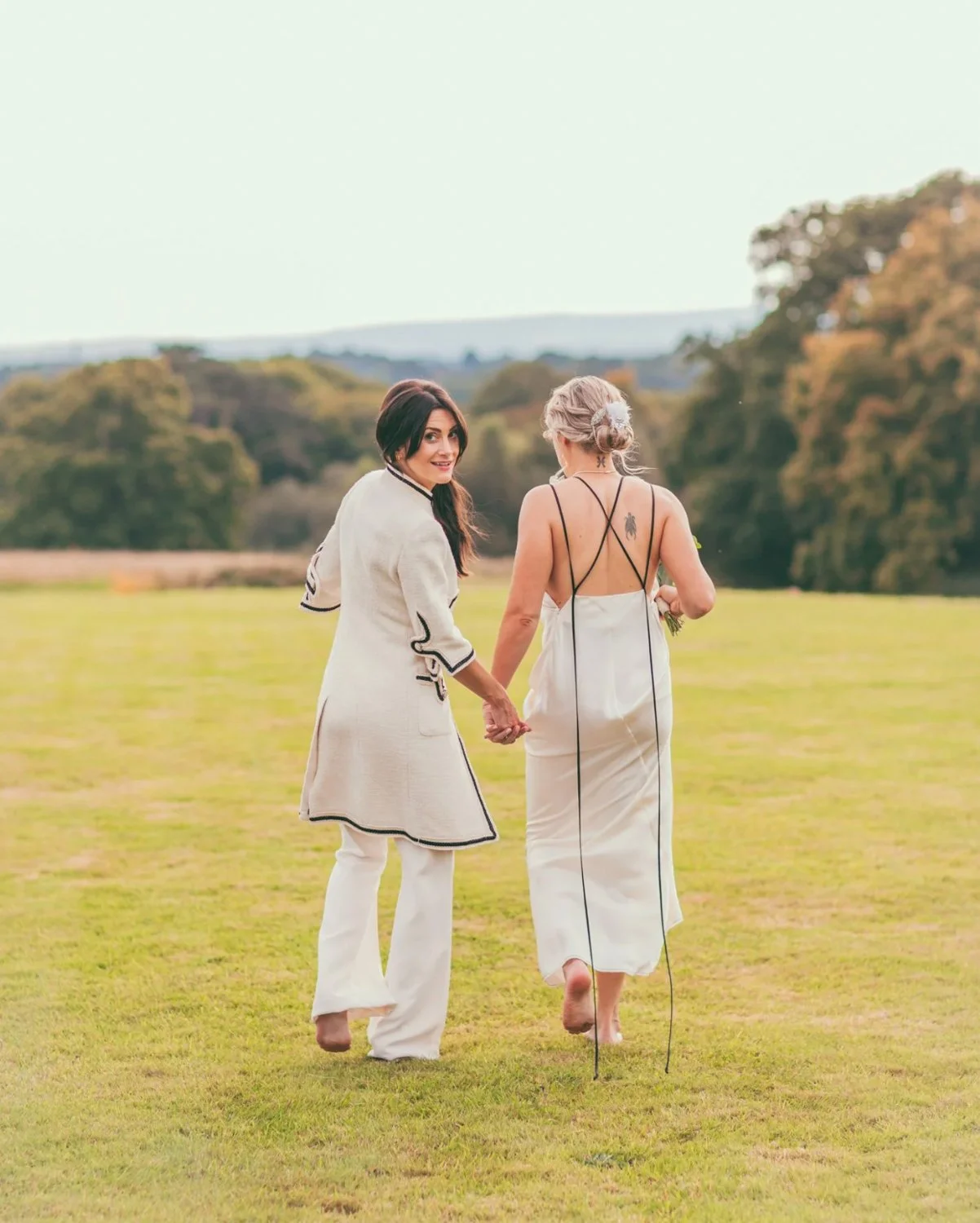 Two women in white dresses holding hands and walking in an open grassy field with trees in the background.