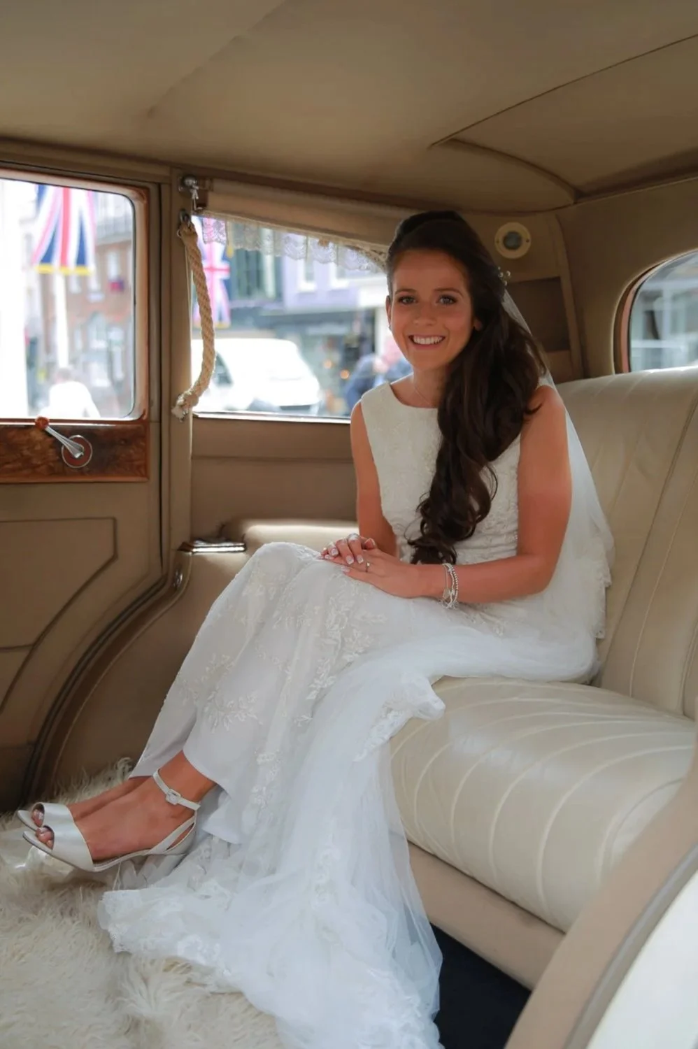 A woman in a wedding dress sitting inside a vintage car, smiling at the camera.
