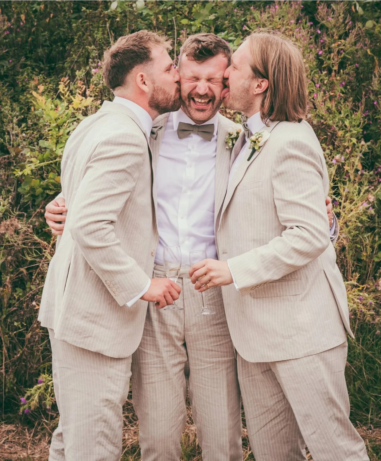 Two grooms kissing the man in the middle on the cheeks during a wedding celebration outdoors.
