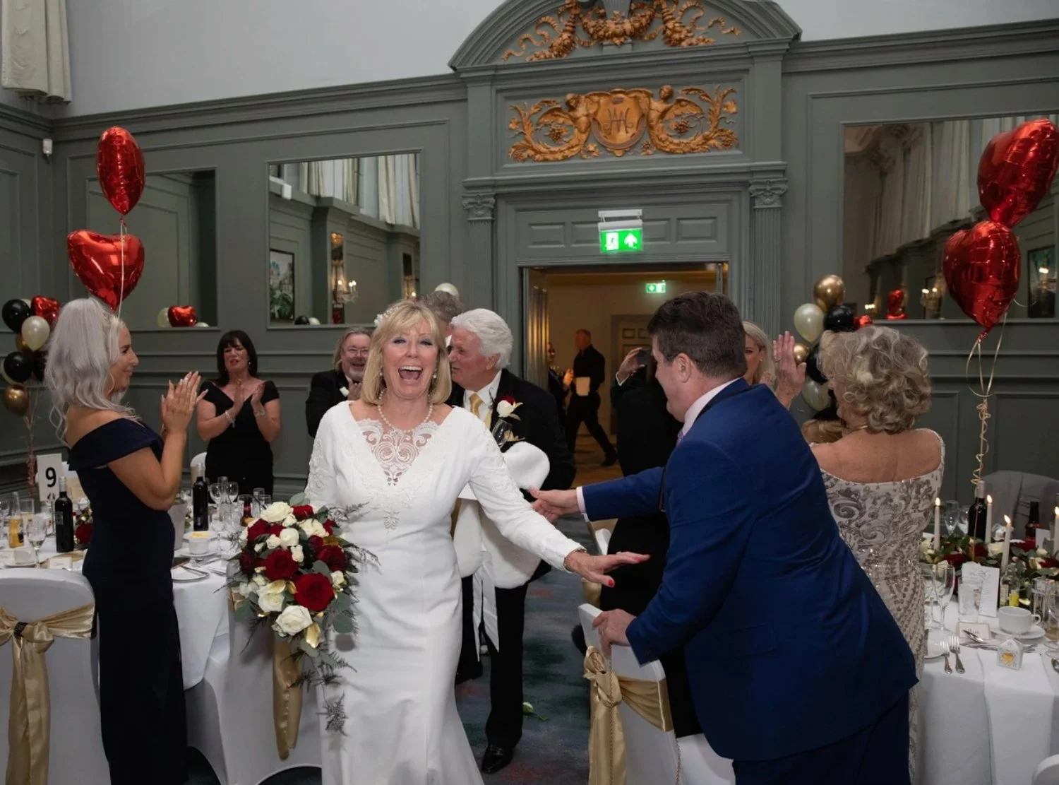 A wedding celebration with a bride in a white dress and a groom in a blue suit, smiling and exchanging vows under grey walls with gold accents, surrounded by guests clapping and balloons.