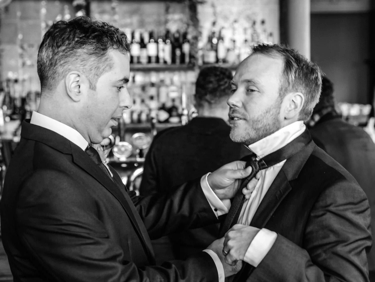 Two men in tuxedos adjusting ties in a bar or restaurant setting, with a bartender and bar shelves in the background.