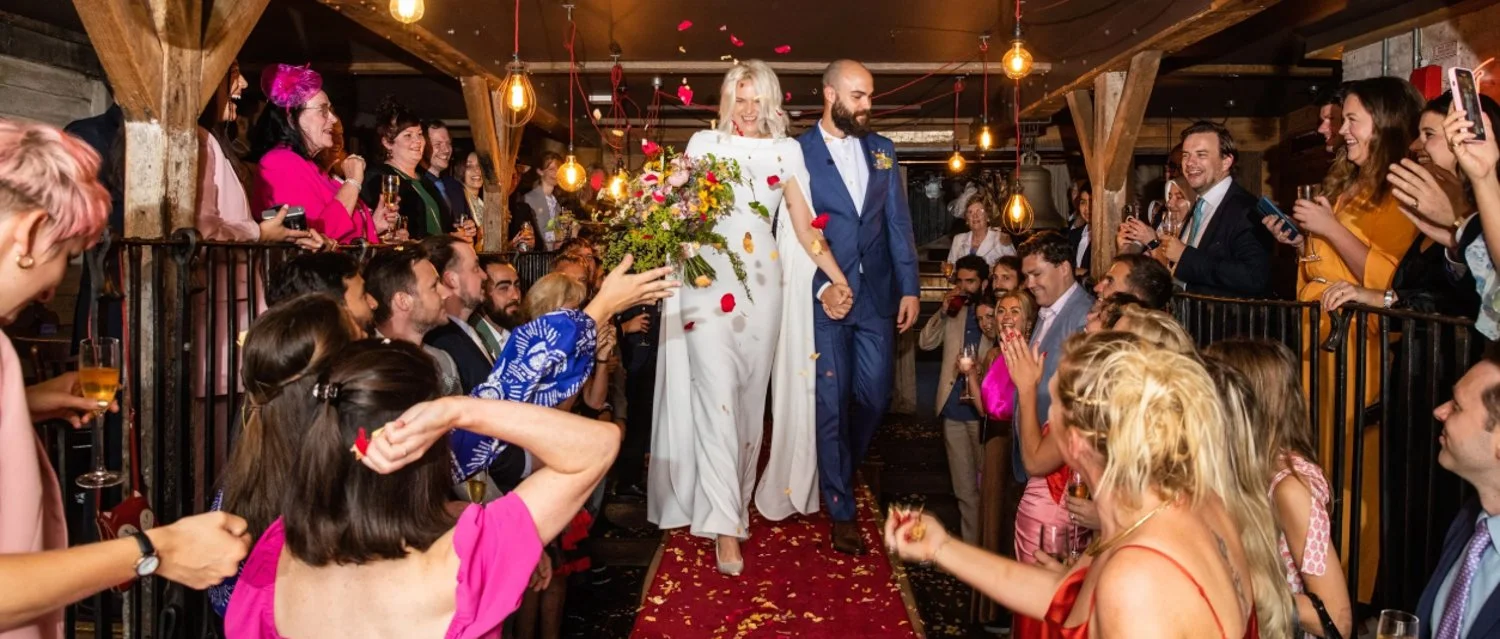 Bride and groom walking down the aisle at their wedding reception, surrounded by guests taking photos and celebrating, with flower petals falling around them.