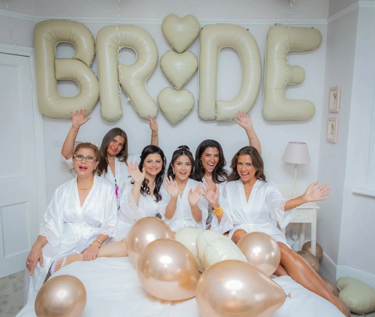Six women in white robes gathered on a bed decorated with golden balloons for a bridal shower, with large helium balloons spelling 'BRIDE' and heart-shaped balloons in the background.