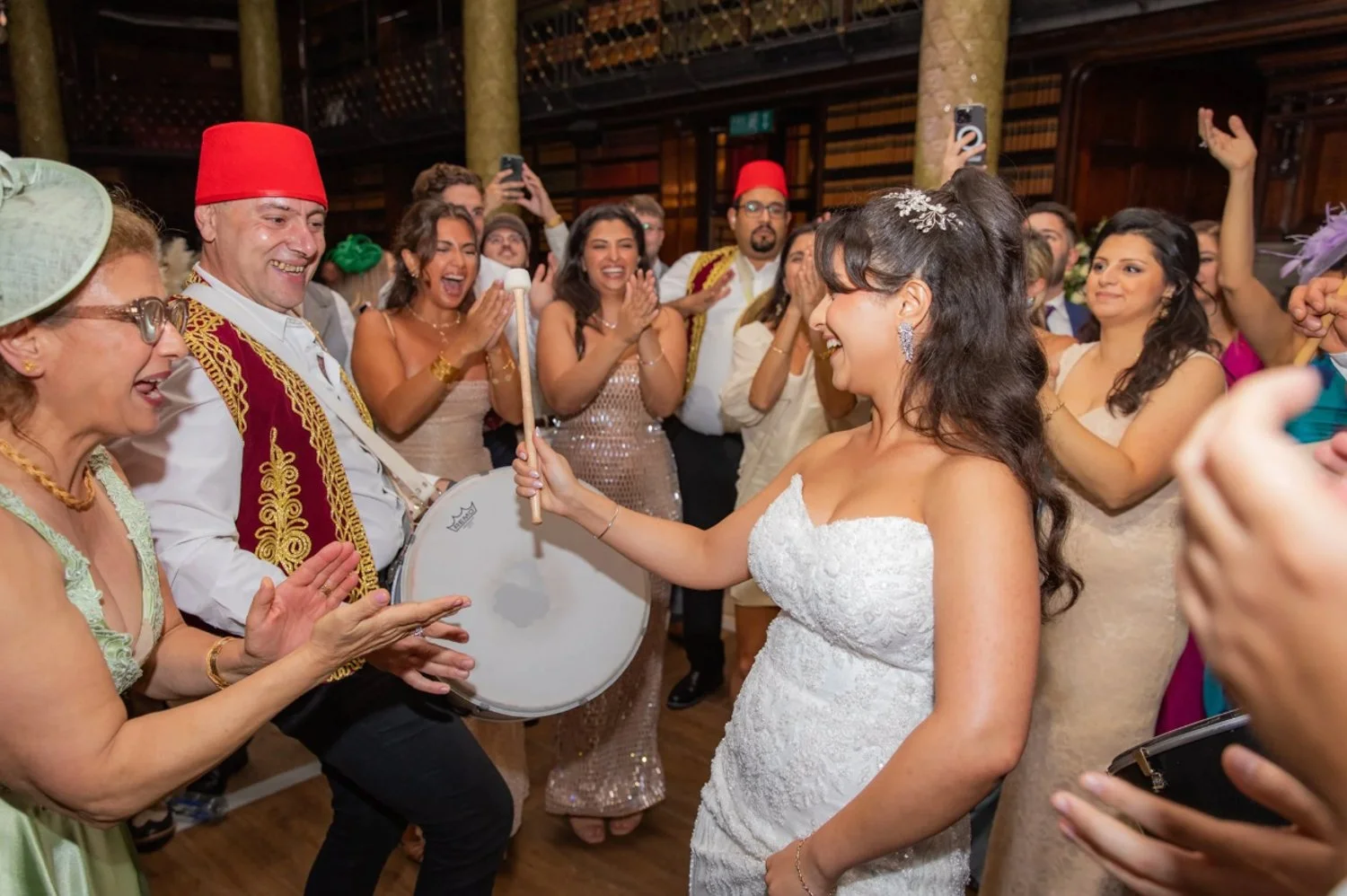 A bride in a white wedding dress smiling while holding a drum stick during her wedding celebration, surrounded by family and friends in festive attire.