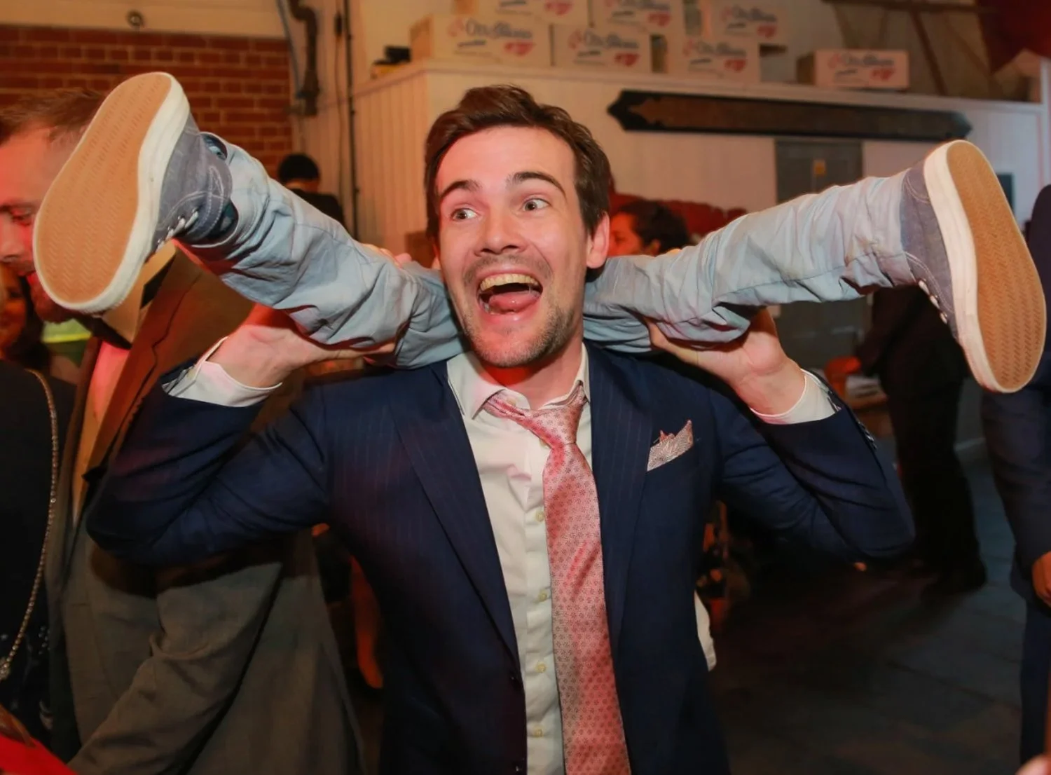 A man in a suit and tie carries a young boy on his shoulders at a lively indoor event, with people in the background.