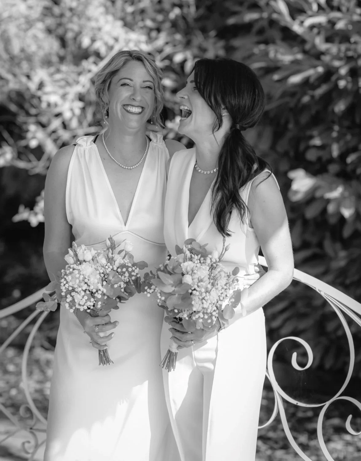 Two women in white dresses holding bouquets, smiling and laughing, outdoors with leafy background.