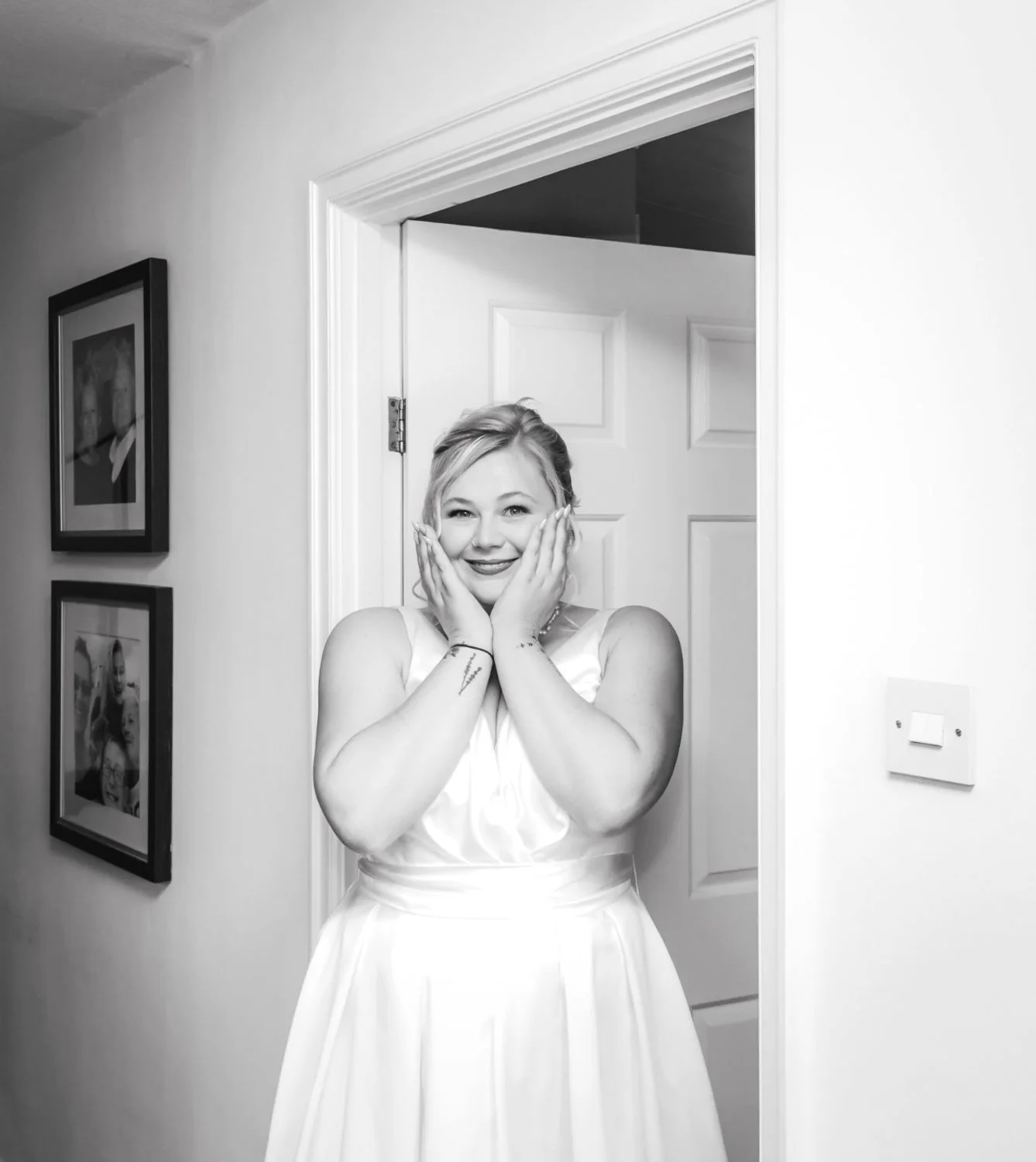 Black and white photo of a smiling woman in a dress, standing in a doorway with her hands on her face, inside a room with framed pictures on the wall.