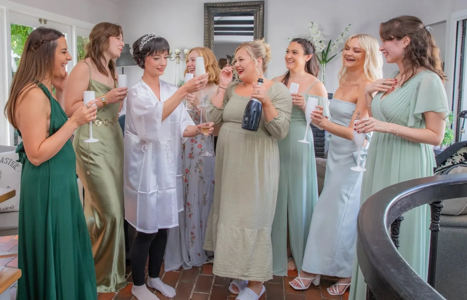 A group of women celebrating together indoors, dressed in pastel-colored dresses and holding champagne glasses, with one woman pouring champagne from a bottle.