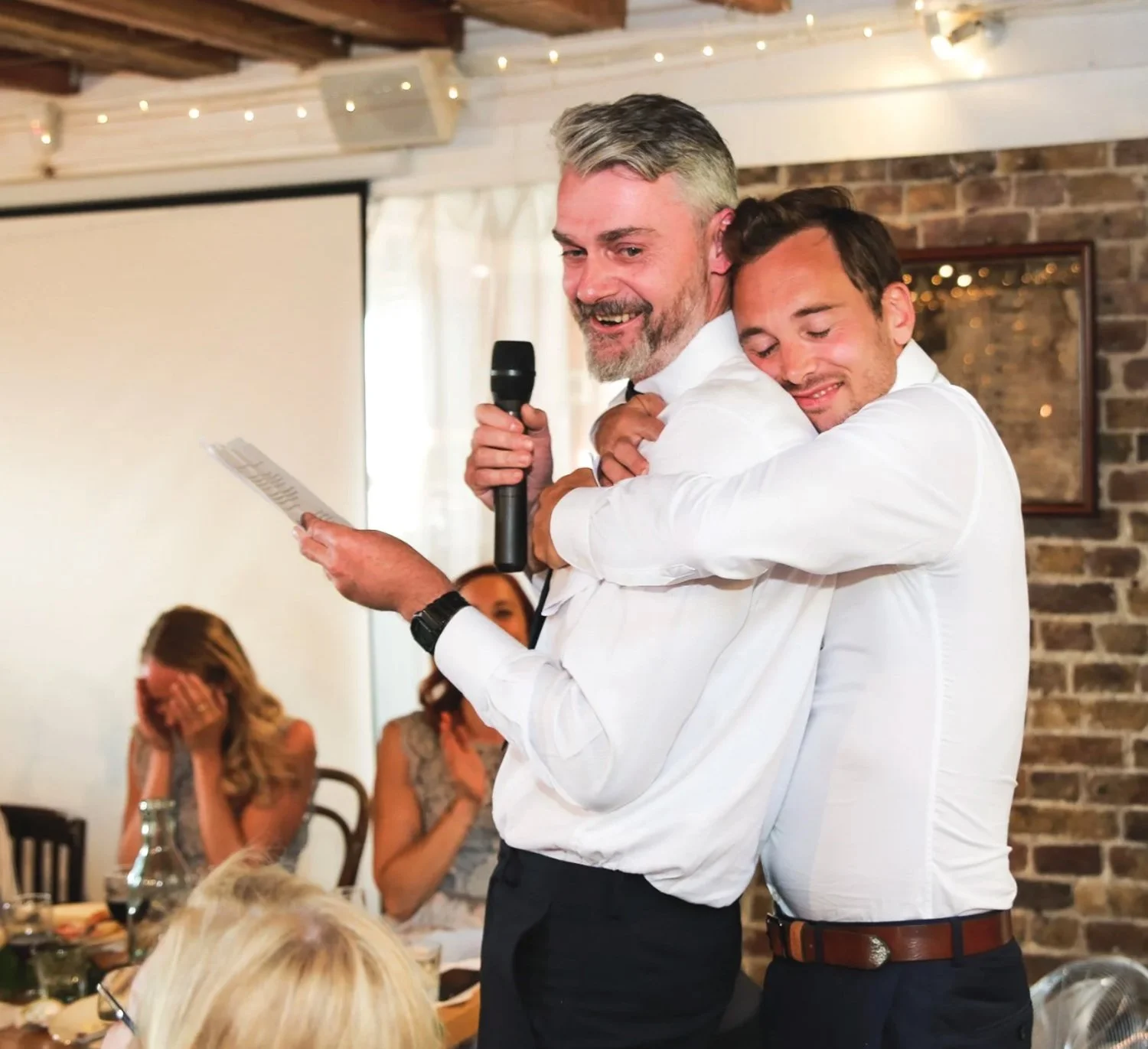 Two men in white shirts hugging, one holding a microphone and reading from a paper, at a celebration with guests seated at a table in the background.