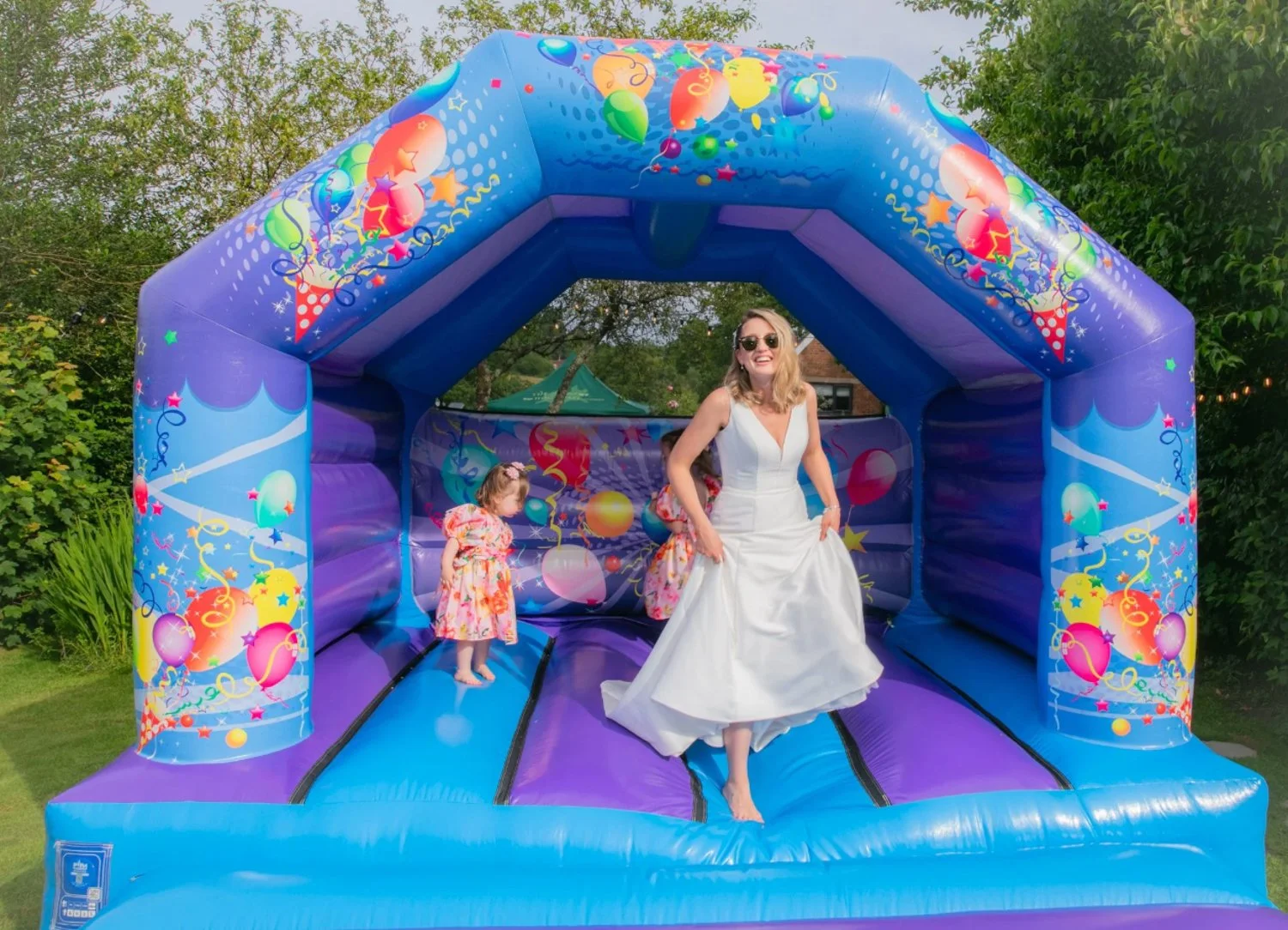A woman in a white dress and sunglasses stands on an inflatable bounce house decorated with colorful balloons and streamers. A young girl in a floral dress stands nearby inside the bounce house. The setting is outdoors with green trees and bushes.