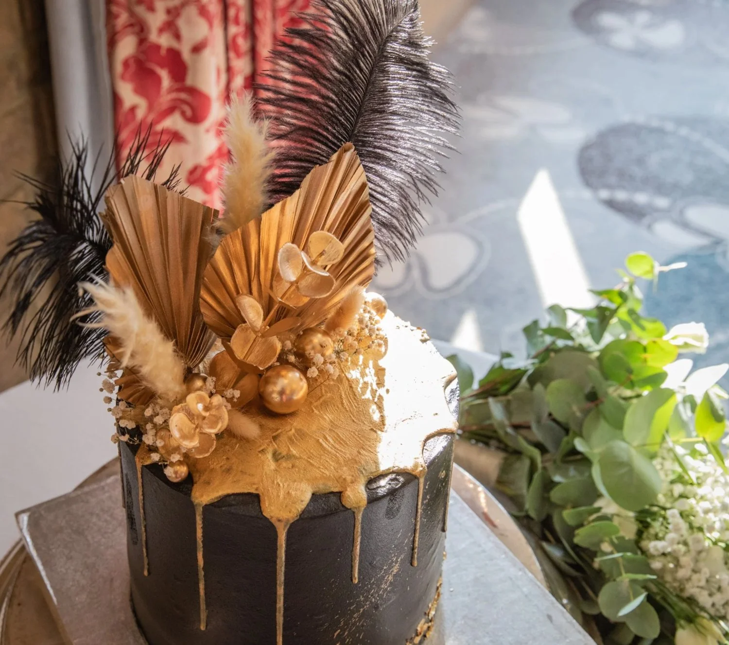 A black and gold cake with gold drip decoration, topped with dried flowers, leaves, and berries. A bouquet of greenery is beside the cake.