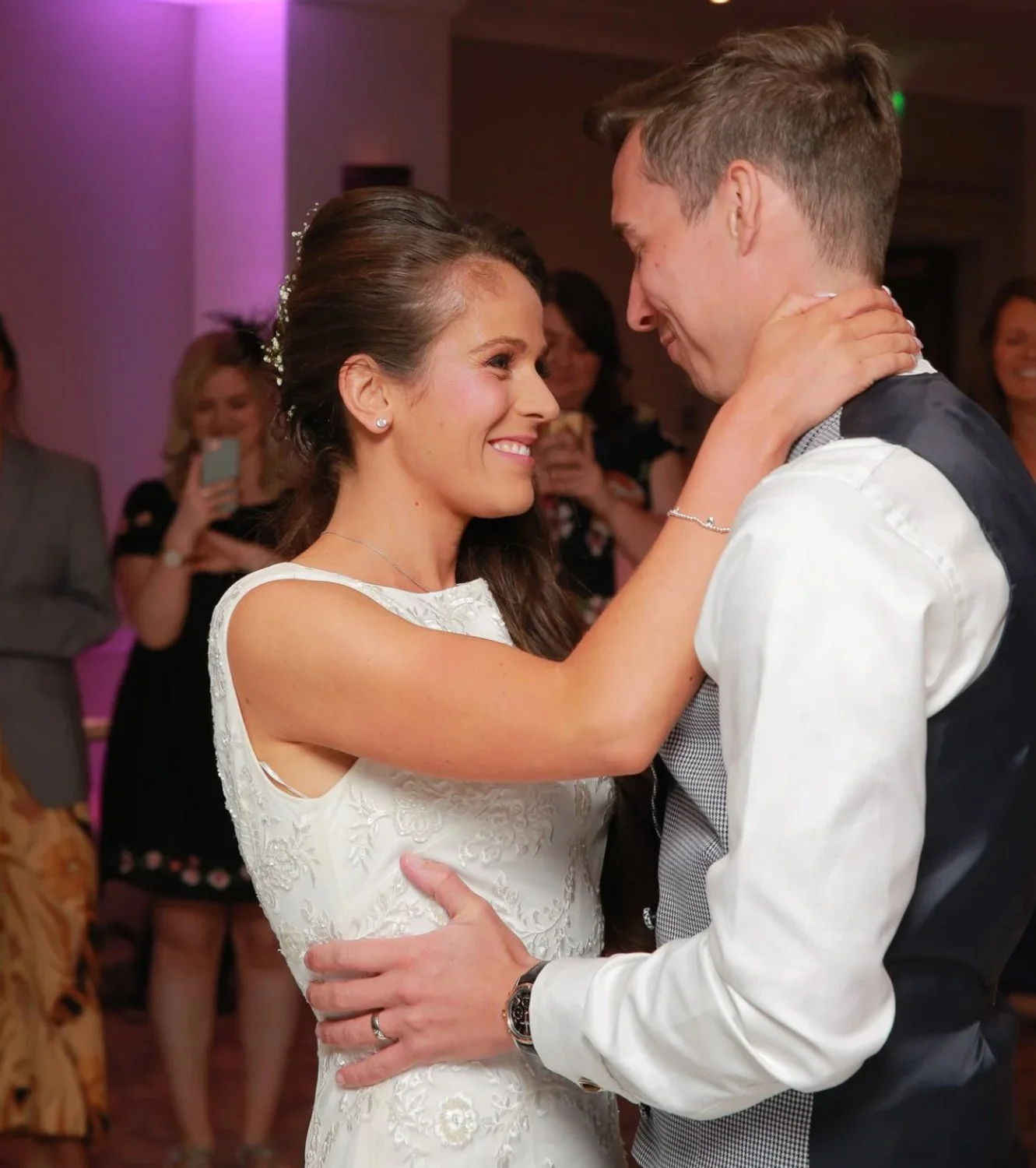 Couple dancing at their wedding reception, smiling and looking into each other's eyes with friends in the background.