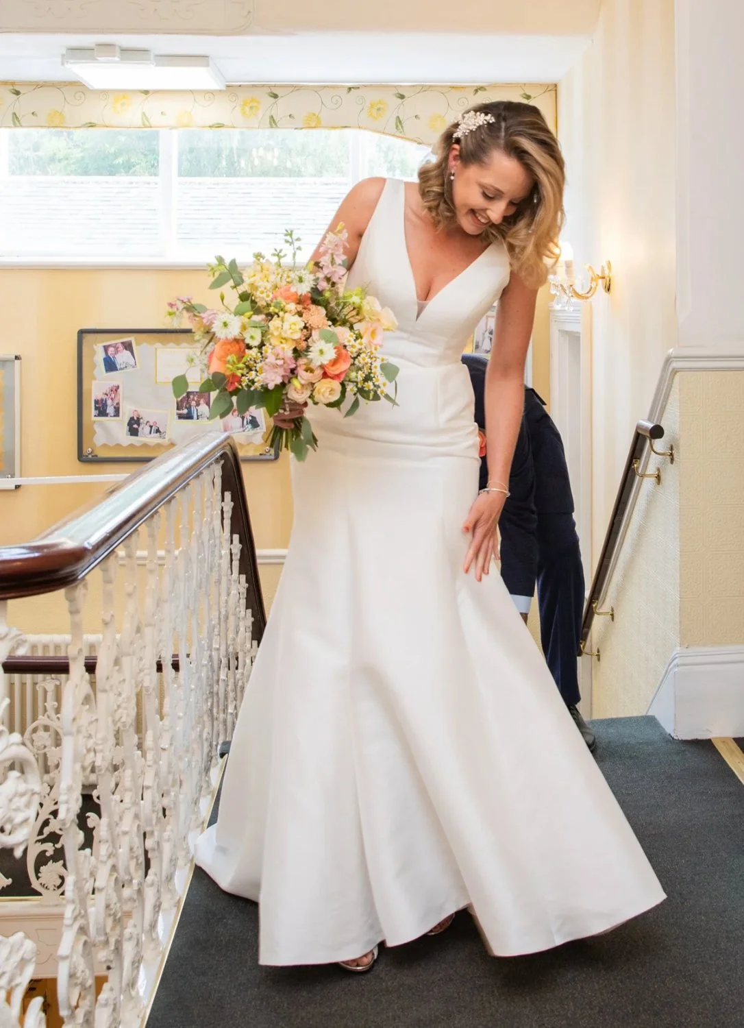 A bride in a white wedding gown holding a bouquet of pink, yellow, and white flowers, standing on an indoor staircase shortly before her wedding, smiling and looking down.